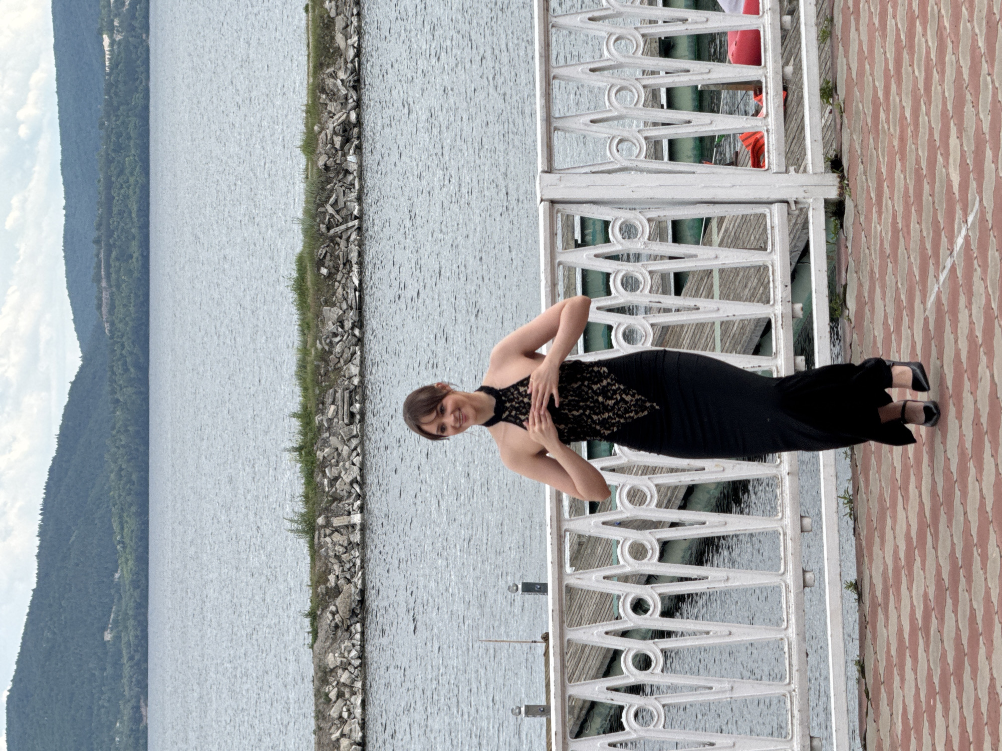 The image displays a full-length portrait of a female subject in an outdoor waterfront setting, identified as Unknown, Unknown.
In the foreground, a paved surface with a red and light-brown checkered pattern extends from the bottom of the frame. Immediately behind this pavement is a white, decorative metal railing featuring circular and vertical bar elements, separating the paved area from a body of water.
The central subject is a young adult female, positioned slightly right of the frame's center. She wears a floor-length, form-fitting black gown with a halter neckline. The bodice of the dress features a prominent black lace overlay over a lighter lining, creating a contrasting pattern across her chest. Her dark hair is styled with bangs and appears pulled back. She is smiling broadly with an open mouth, looking slightly to her left. Her hands are clasped together at chest height, resting on the lace detail of her dress. She wears black high-heeled shoes.
Behind the railing, a wooden dock structure is visible on the calm, grey-blue water. Partially obscured objects with green and red coloration, possibly boats or kayaks, are visible behind the railing and dock. Further into the midground, a rocky breakwater or land formation covered with sparse vegetation stretches horizontally across the water. The background features a large expanse of water, leading to a distant shoreline dominated by heavily forested hills and mountains under a partly cloudy sky. The lighting suggests daytime conditions.