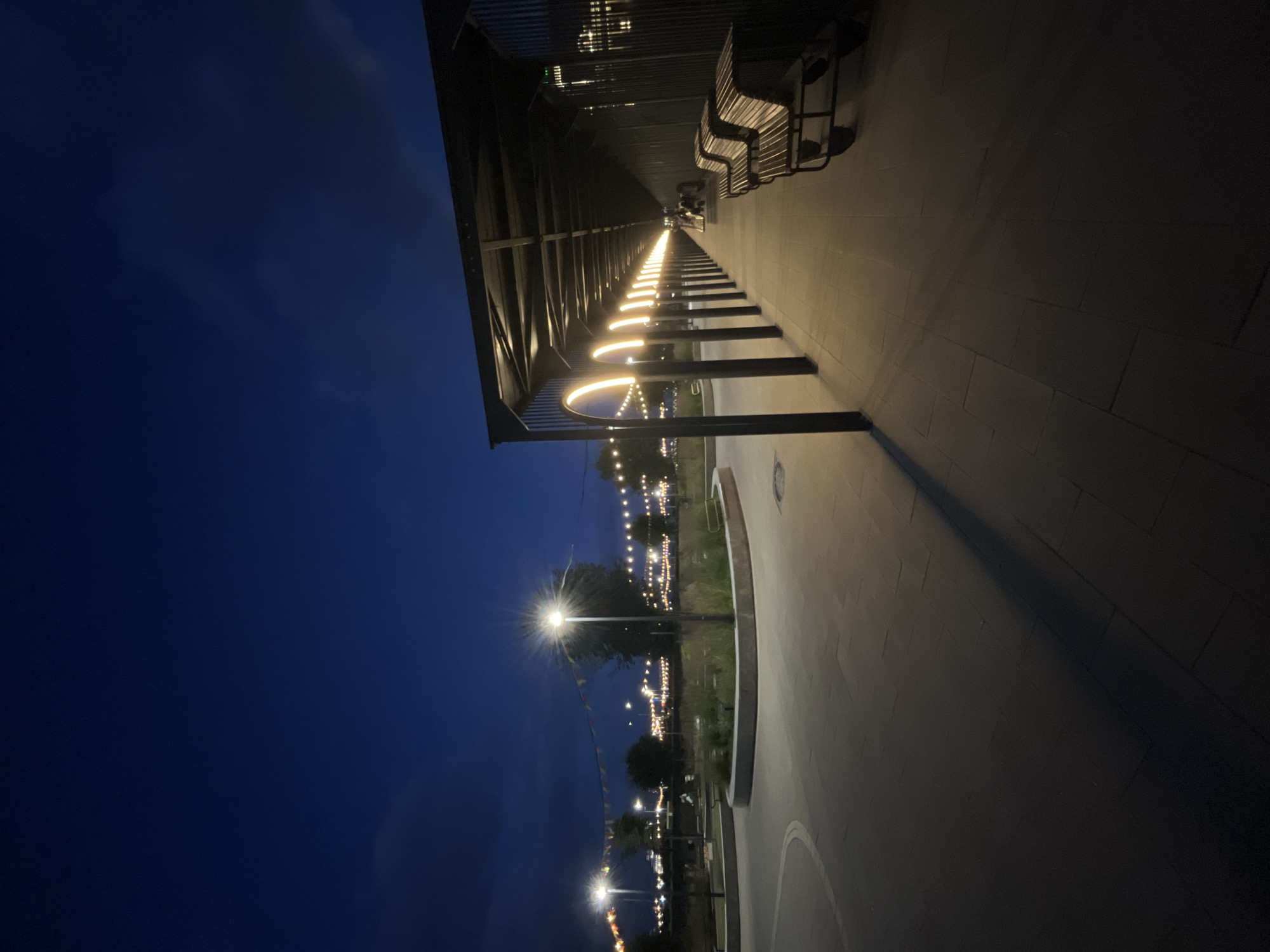 The image captures a pathway at dusk or early night in an urban park setting in Nizhny Novgorod, Russia. The sky is a deep navy blue with some wispy clouds. On the right side of the image, a covered walkway with a corrugated metal roof extends into the distance, supported by pillars. Along this walkway, semi-circular lights are embedded in the ceiling, casting a warm glow on the paved ground. Two park benches are visible under this structure, made of wooden slats and metal frames.
To the left of the walkway, a paved pathway curves and leads into the park. In the middle ground, a raised circular garden bed contains greenery and a lamppost that emits a bright light, creating a starburst effect. Further into the park, strings of festive lights are draped between trees and poles, illuminating the landscape. The overall atmosphere is serene and inviting, suggesting a public space designed for evening recreation or events.