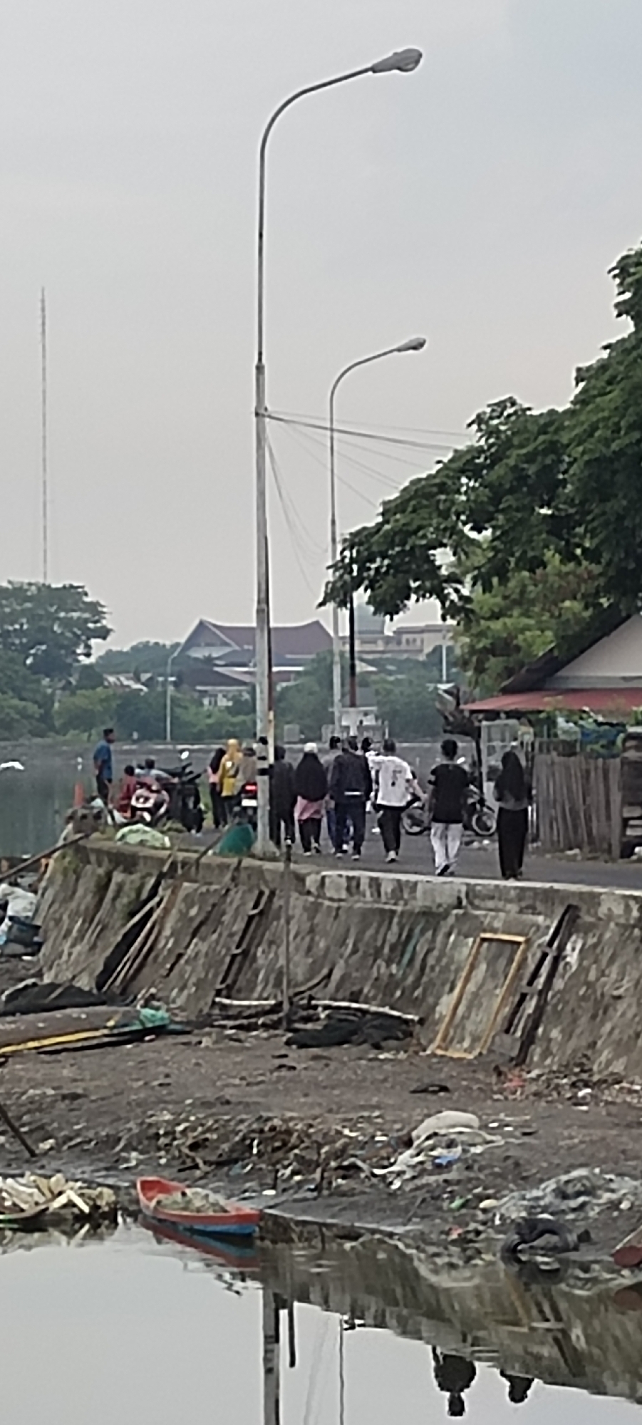 The image shows a waterfront scene in Lhokseumawe, Indonesia, with a focus on a concrete embankment littered with debris and a shallow body of water in the foreground. Several people are walking on the road adjacent to the embankment, some with motorcycles. The background reveals trees, buildings, and streetlights. The overall impression is of a somewhat neglected urban environment.