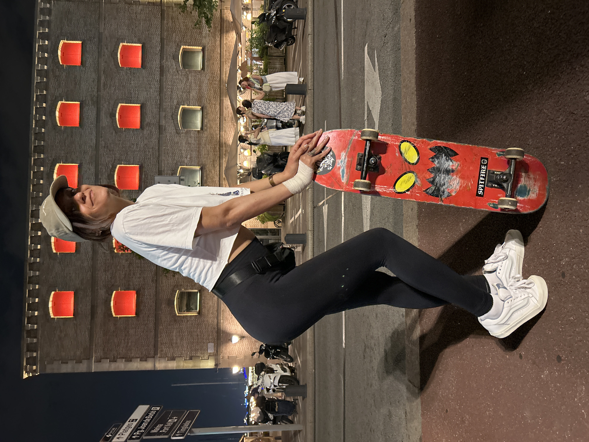 A young woman poses with a red skateboard on a city street at night. She wears a cap, a cropped white t-shirt, black leggings, and white sneakers. Her hands are wrapped in what appears to be bandages. The skateboard features a "Spitfire" sticker and faded graphics. Behind her is a tall brick building with multiple windows illuminated in red, and street signs in the lower left corner suggest the location is in Barcelona, Spain. Other people and parked motorcycles are visible in the background along the street. The composition is tilted slightly to the right.