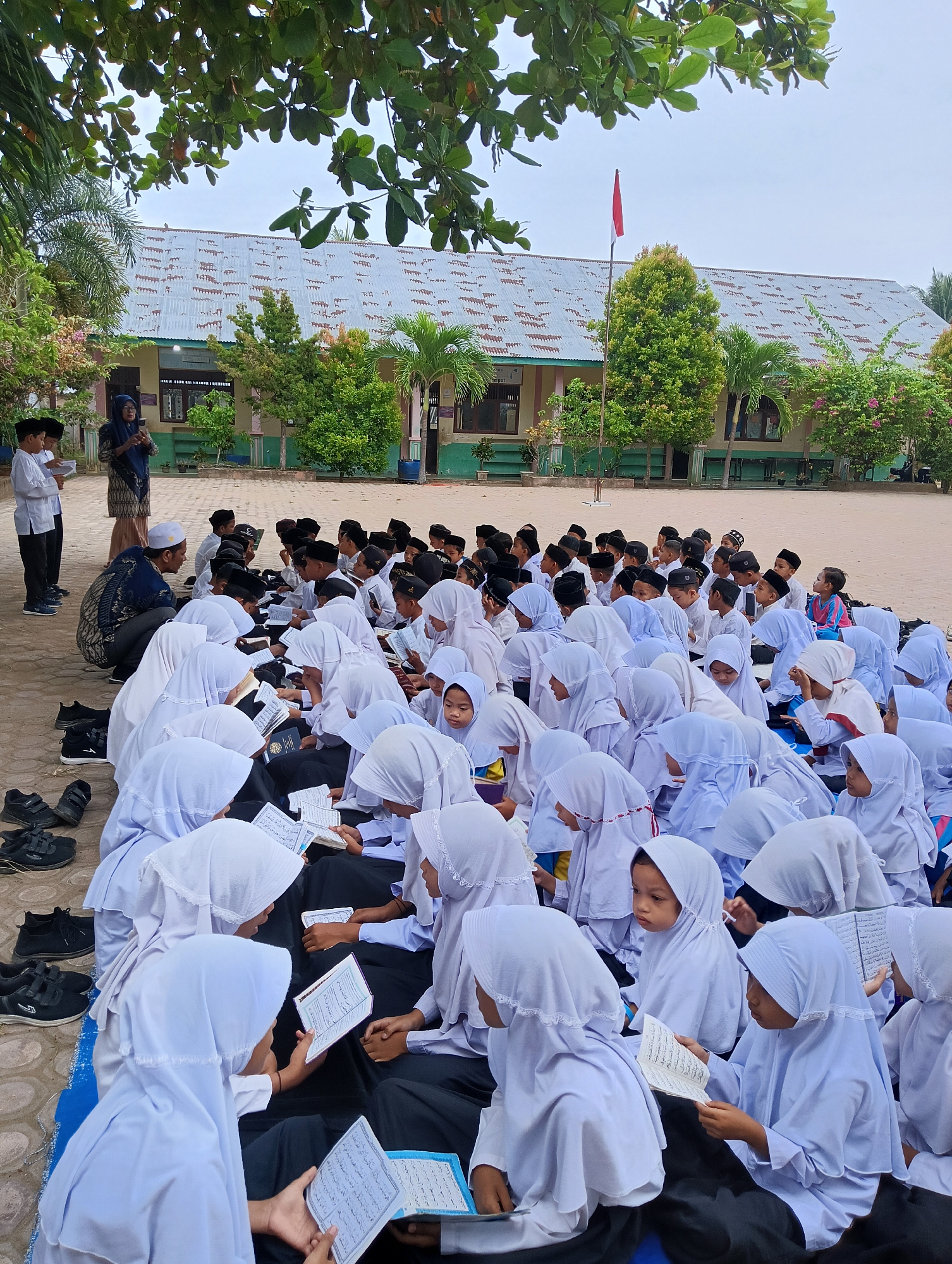 In Baratpayaitek, Indonesia, a group of Muslim schoolchildren are gathered outdoors, seemingly for a lesson or prayer. The children, dressed in traditional white Islamic attire, including hijabs for the girls and peci (caps) for the boys, are seated on the ground, facing a woman who appears to be an instructor. Many of the children are holding and reading from religious books, suggesting a religious education session. The setting appears to be a schoolyard, with a building in the background and trees providing shade. Several pairs of black shoes are neatly placed on the ground near the edge of the gathering. The atmosphere seems peaceful and orderly.