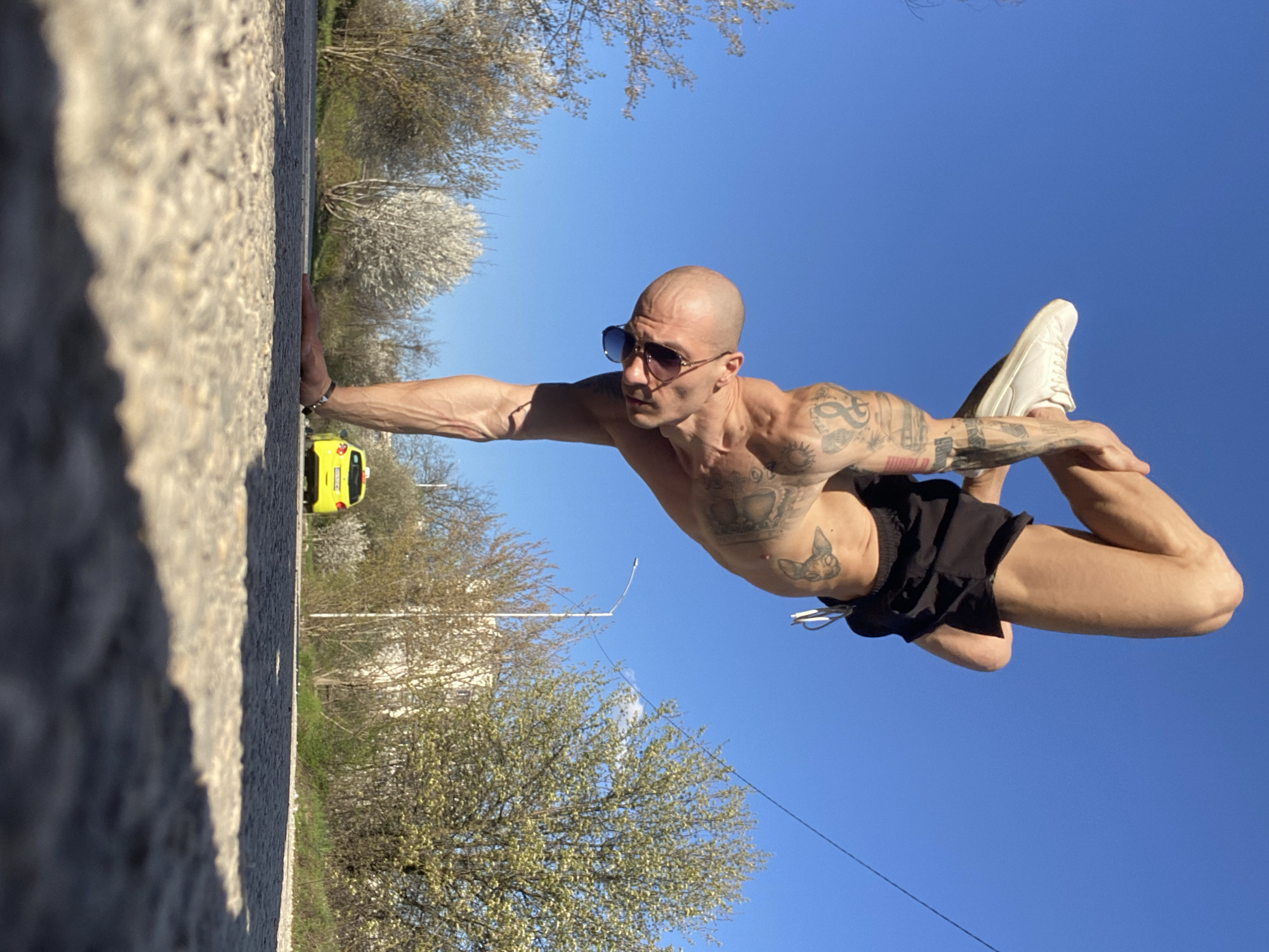 A bald, muscular man wearing sunglasses and black shorts is performing a yoga pose on the edge of a curb next to a street. He is balancing on one hand pressed against the curb, his body extended horizontally, and the other hand holding his bent leg in the air. He is wearing a white shoe on the foot of the bent leg. His torso is bare, revealing several tattoos. In the background, there is a yellow car parked on the street, trees with white blossoms, and a building. A utility pole and wires are visible against the blue sky. The location is in Pleven, Bulgaria.