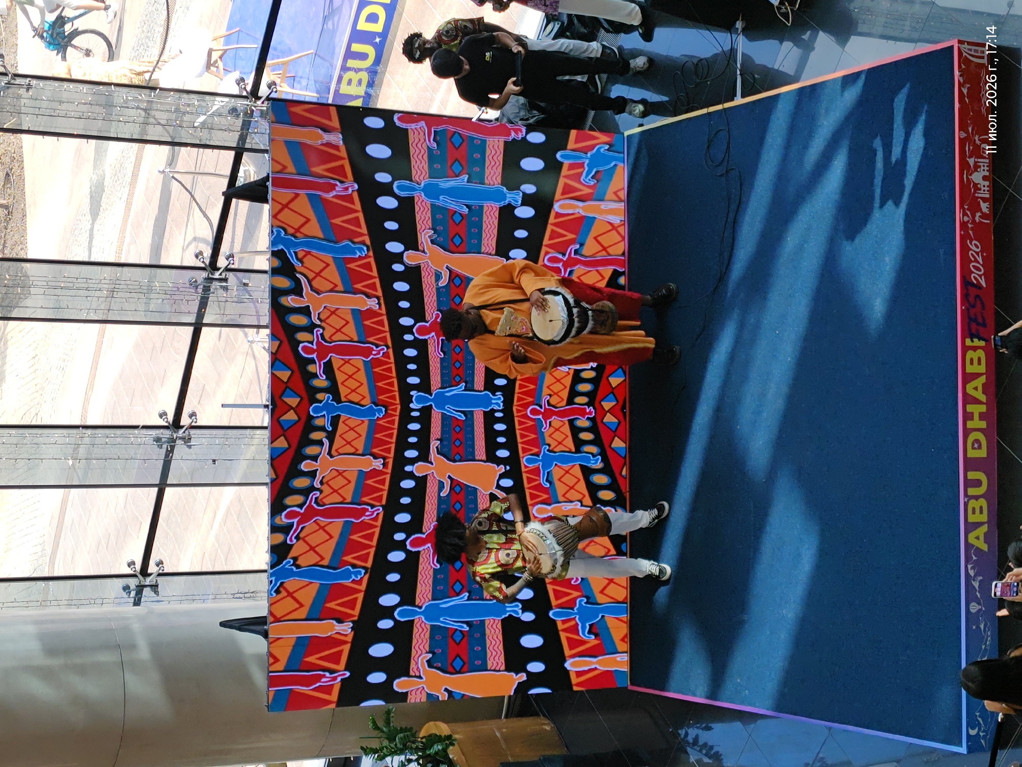 The image depicts a lively outdoor event with a stage set up in front of a large, colorful backdrop featuring abstract patterns and stylized figures in vibrant reds, blues, and oranges. Two individuals are performing on the stage. One person, dressed in traditional African attire with a patterned shirt and white pants, is playing a drum strapped to their waist. Another person, wearing an orange robe, is also playing a drum. To the right of the stage, several people are observing the performance. One individual in a black t-shirt and pants is holding a tablet, possibly recording or managing the event. The background reveals an urban setting with modern architecture, including glass facades and metal structures, suggestive of a public space like a plaza or a cultural center. A bicycle is visible in the distance, adding to the sense of an active city environment. The overall atmosphere appears festive and engaging, with a focus on cultural performance. The text on the bottom of the stage banner reads "ABUFEST 2026" and "11 июл. 2026 г., 17:14". The event is located in Astana, Kazakhstan.