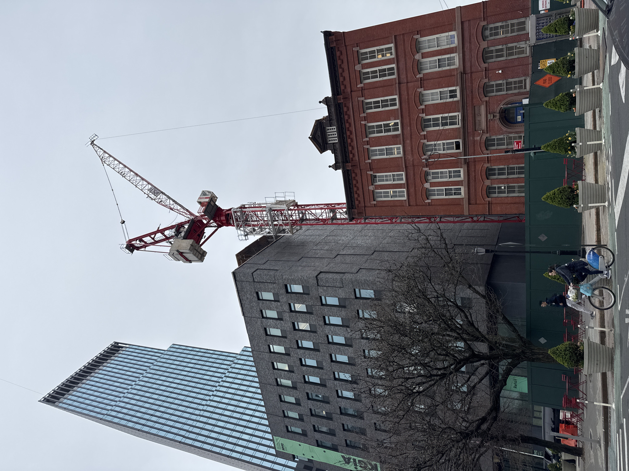 The image shows a street scene in New York City, United States of America. A red and white construction crane dominates the view, positioned between several buildings. To the left is a modern glass-fronted skyscraper. To the right is a red brick building, and in the center, a grey building. A leafless tree is in the foreground. On the right side of the image, two people are riding bicycles. A green construction fence runs parallel to the street, with decorative plants in pots placed along the sidewalk.