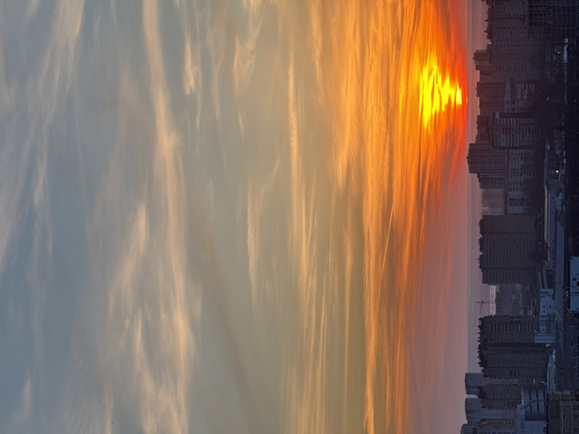 The image displays an elevated panoramic view of the city of Perm, Russia, featuring an urban expanse including what appears to be the Kama River, captured during either sunset or sunrise. The sky, occupying the upper two-thirds of the frame, exhibits a gradient of colors, transitioning from light blue-grey at the top to hues of peach, orange, and intense red at the horizon. Wispy, horizontally oriented clouds are distributed across the sky, becoming more defined and colorful towards the lower portion. A distinct, bright yellow-orange sun, horizontally elongated and somewhat distorted by atmospheric conditions, is positioned centrally near the horizon line. A faint white contrail extends diagonally in the upper right quadrant.
The lower third of the image depicts a dense urban landscape. Numerous multi-story buildings, predominantly appearing as dark silhouettes against the bright sky, comprise the foreground and mid-ground. These structures vary in height and density, characteristic of residential and commercial blocks. A wide, dark body of water, consistent with the Kama River, is visible beyond an immediate cluster of buildings, stretching horizontally across the middle distance. A large construction crane is discernible in the mid-ground, slightly to the left of the sun's alignment, indicating ongoing development. The distant urban horizon appears hazy and less defined. No discernible individuals, specific actions, or interactions are visible within the frame.