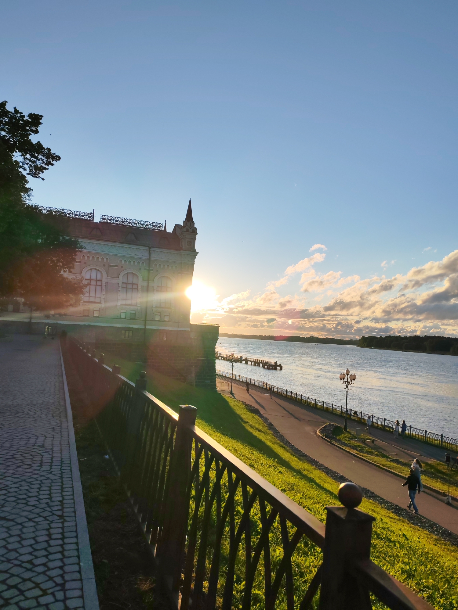This image captures a sunset view along a waterfront promenade in the city of Bol'shie Mkhi, Russia. In the foreground, a dark metal fence borders a cobblestone pathway and a grassy embankment. Beyond the fence, a paved walkway curves along the edge of a wide river. Several lampposts with decorative tops are spaced along this walkway, where a few individuals are seen walking and cycling. On the left side of the image, a large, ornate building with arched windows and a pointed spire stands prominently, partially obscured by a tree in the foreground. The setting sun is visible just behind the building, casting a warm glow and lens flare across the scene. The sky above is a clear blue with scattered, fluffy white clouds illuminated by the sunset. The river stretches to the horizon, where a tree-lined shore is visible.