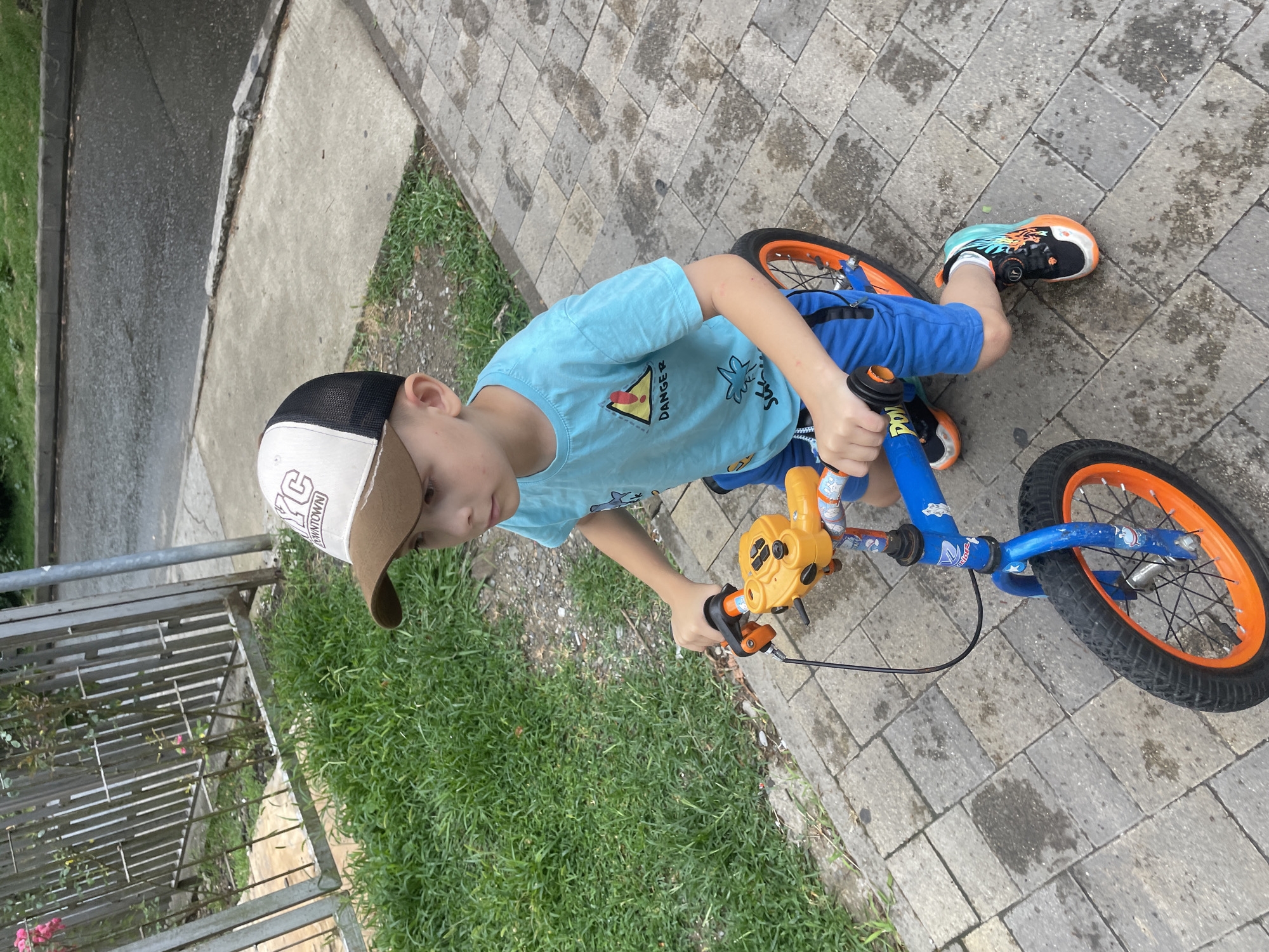 A young boy with fair skin, short brown hair, and brown eyes is wearing a light blue t-shirt with a "DANGER" graphic and blue shorts. He is also wearing a brown and white baseball cap and multi-colored athletic shoes. The boy is seated on a blue balance bike with orange accents, holding the handlebars, and appears to be looking to his right. The bike has an orange front wheel. He is on a paved walkway made of grey interlocking pavers, which are wet in patches, suggesting recent rain. To the left of the walkway, there is a patch of green grass and a metal fence with a rose bush growing through it. Behind the fence, there is a grey concrete curb and a dark asphalt road. The setting appears to be an outdoor area, possibly a park or residential street in Sochi, Russia, given the characteristic paved walkways and vegetation. The overall lighting suggests it is daytime.
