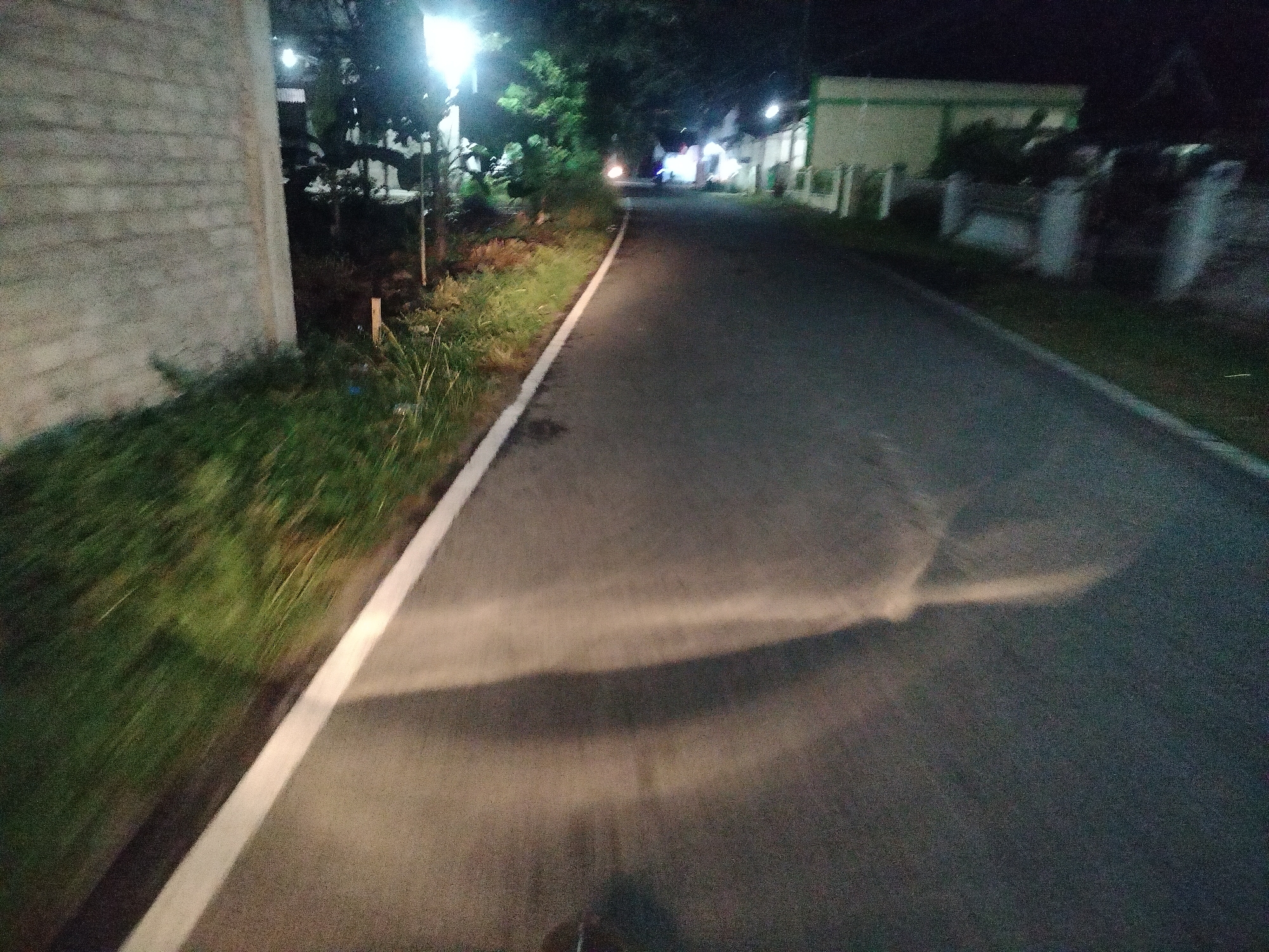 The image depicts a nighttime street scene in Kumbangindah, Indonesia. A paved road runs through the center of the frame, bordered by a grassy area on the left and buildings on the right. A white line runs along the edge of the road, separating it from the grass. Streetlights illuminate the scene, casting shadows on the road. The buildings appear to be low-rise residential or commercial structures. The overall impression is one of a quiet, dimly lit street at night.