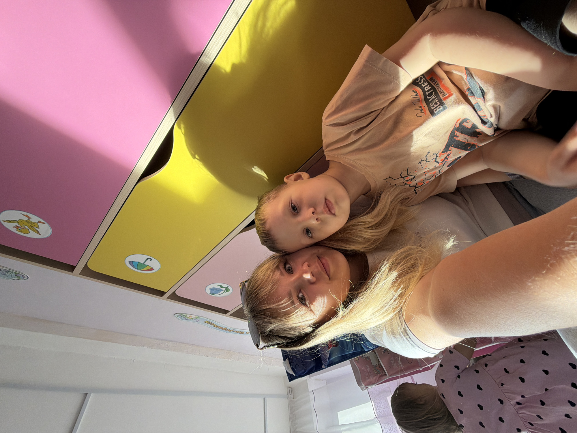 A selfie, taken at an unusual angle, appears to show a woman and a young child in an indoor setting. The woman has blonde hair and is wearing sunglasses on top of her head. The child, also with blonde hair, is wearing a t-shirt with a graphic design. They are positioned in front of a wall unit featuring colorful drawers, with pink and yellow panels visible. The ceiling is visible above, with decorative stickers affixed to some of the drawers. The location might be in the city of Chaikovsky, Russia, based on the instructions, although this is not directly evident from the image itself.