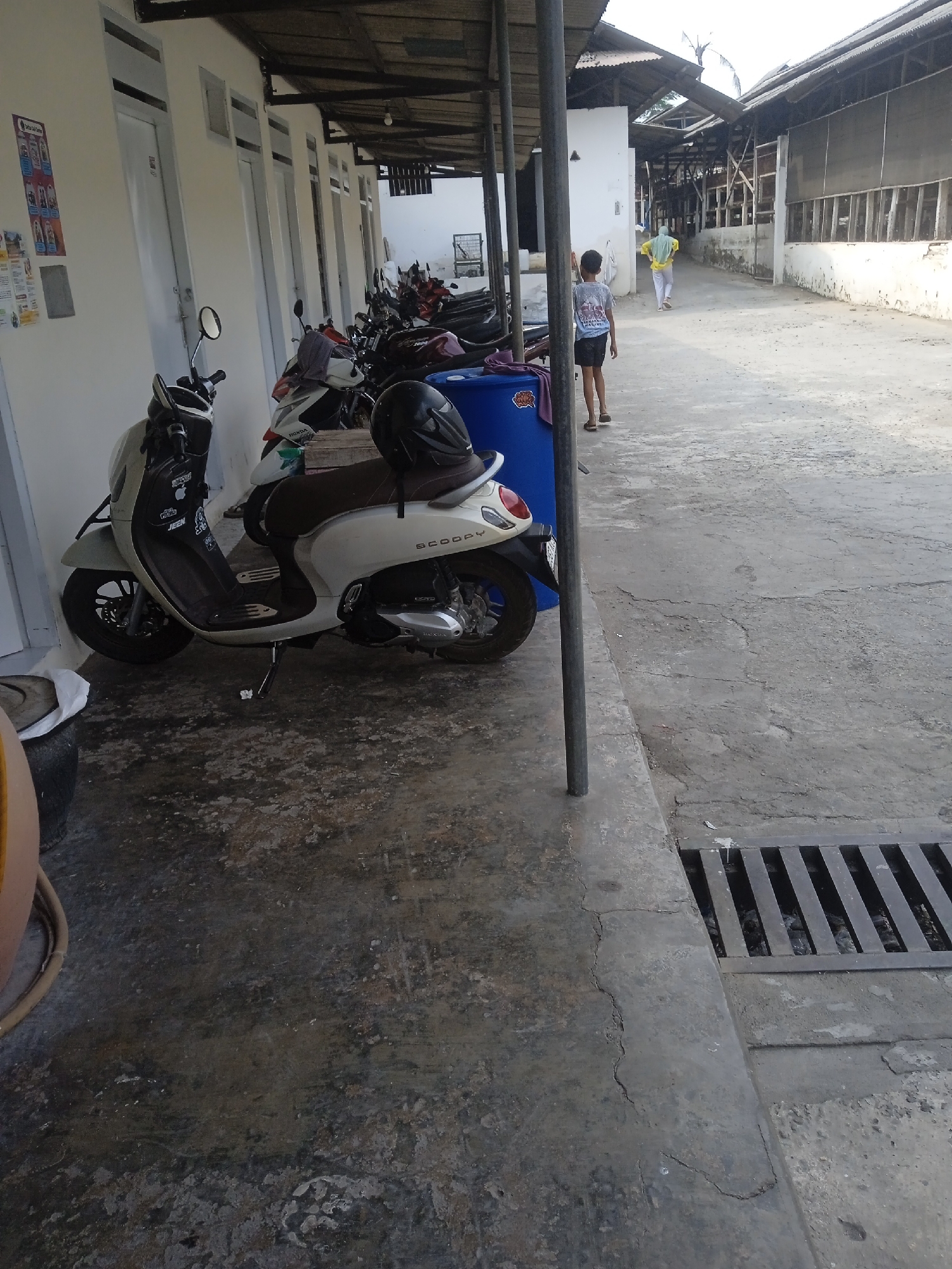 This image depicts an outdoor scene in Slorok, Indonesia, possibly near residential or commercial buildings. In the foreground, a light-colored scooter is parked, with a black helmet resting on its seat. The scooter is adorned with stickers. To the left, a portion of a yellow barrel is visible. Behind the scooter, several other motorcycles are parked in a row.
The ground is paved with concrete that shows signs of wear and cracking. A metal grate, likely a drain cover, is present in the lower right portion of the image. In the background, two individuals are walking away from the camera down a pathway. One person is wearing a white shirt and dark shorts, while the other is in a yellow headscarf and white pants. Buildings with corrugated metal roofs line the pathway, suggesting an urban or semi-urban environment. The overall impression is one of a functional, possibly residential or small business area.