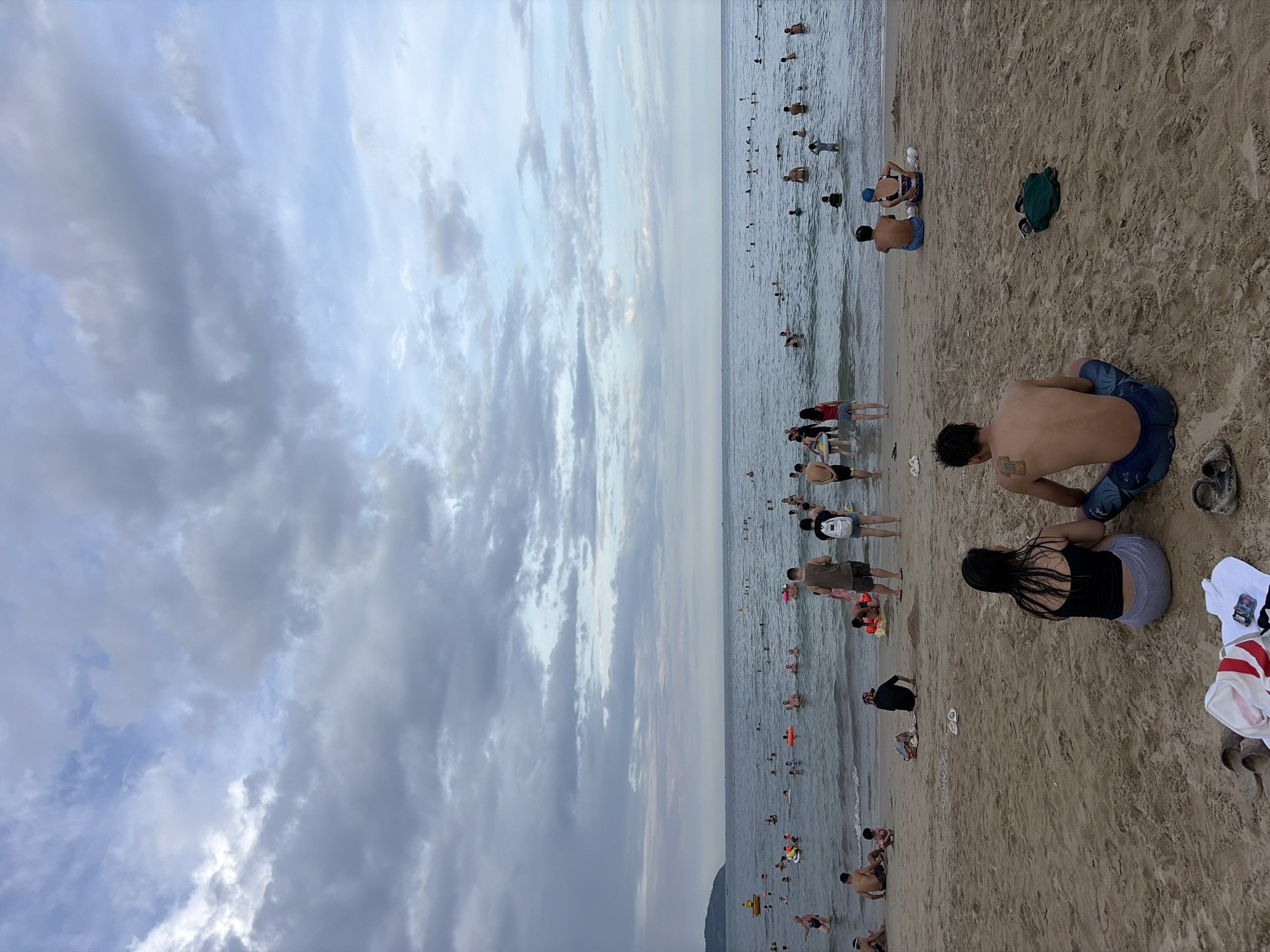 This is a vertical, eye-level, full-shot photograph of a crowded beach scene at what appears to be Mỹ Khê Beach in Đà Nẵng, Vietnam. The sky is overcast with a mix of light and dark gray clouds, with patches of blue sky visible. The ocean is calm, with gentle waves reaching the shore.
In the foreground, two individuals are sitting on the sand, facing away from the camera and towards the water. The person on the left is a woman with dark hair, wearing a black tank top and grey shorts. The person on the right is a man with tattoos on his back, wearing blue swim trunks. Scattered on the sand in front of them are a pair of flip-flops, a white towel with red stripes, and another pair of flip-flops. A dark green bag is placed further to the right.
The middle ground of the image features a sandy beach with numerous people. Several individuals are sitting or standing near the water's edge. Further out, a large number of people are in the ocean, some swimming, others standing in the shallow water, and some are using inflatable rings. A small yellow boat is visible in the distance on the left side of the frame.
In the background, the ocean stretches to the horizon, meeting the cloudy sky. A distant landmass, likely a peninsula or island, is visible on the far left, with some vegetation. The overall impression is a lively beach scene on an overcast day.