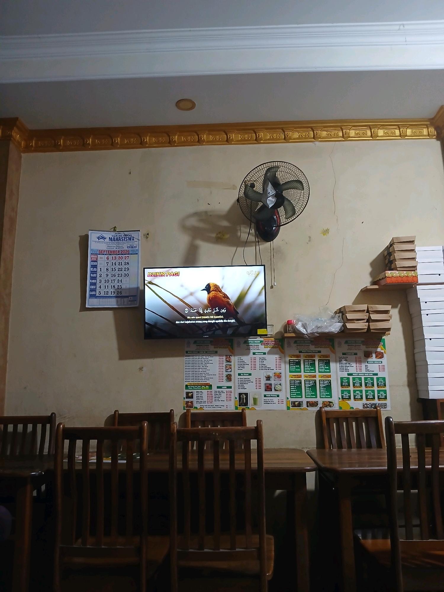 The image depicts the interior of what appears to be a casual dining establishment, possibly a warung or small restaurant, in Depok, Indonesia. The scene is dimly lit, with the primary light source seemingly coming from the television screen and possibly some overhead lighting.
Dominating the wall on the left is a calendar for September 2026, with the header "WARUNG MAHASISWA." Below the calendar, a television screen displays a nature program featuring a bird. To the right of the television, a rotating electric fan is mounted on the wall. Further to the right, shelves are stacked with brown cardboard food containers. Below these shelves and to the right of the television are several laminated menus displayed on the wall, detailing food and drink options with prices listed in Indonesian Rupiah.
In the foreground, several wooden chairs and tables are visible, suggesting a seating area for customers. The overall impression is of a functional, perhaps utilitarian, dining space. The wall appears to be a light beige or cream color and shows some minor imperfections, including a crack running vertically from the fan down towards the television. The ceiling has decorative gold trim. The presence of the "WARUNG MAHASISWA" (Student's Eatery) calendar and menus with local food items strongly indicates the location is in Depok, a city known for its universities.