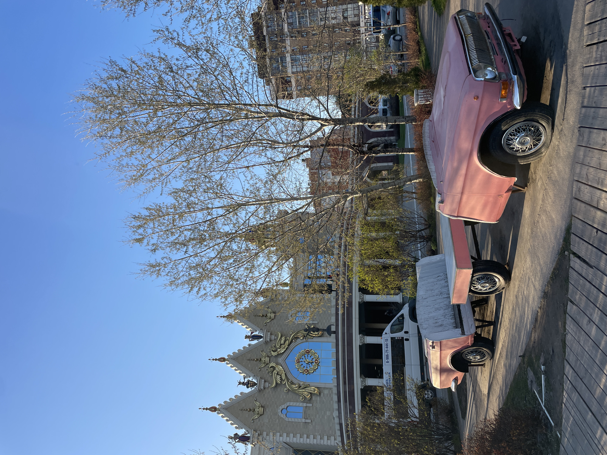 The image shows the Palace of Farmers in Kazan, Russia, viewed from ground level with a clear blue sky above. A bare tree stands in front of the building. To the right of the building, there is a pink vintage car on a flatbed trailer attached to a white van. The image suggests a sunny day in Kazan.