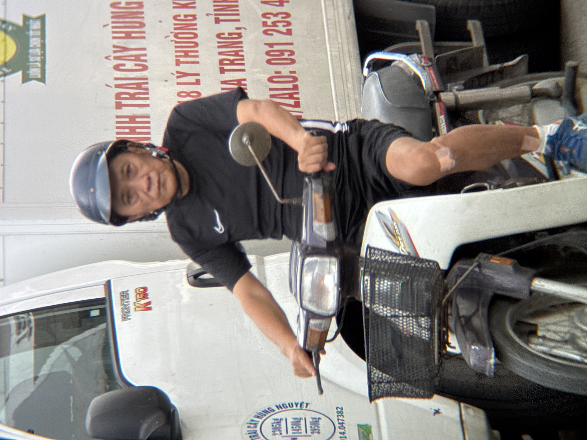 An elderly Asian man wearing a blue helmet and a black Nike t-shirt rides a white motorcycle. The motorcycle has a black basket on the front and a black seat. The man is holding the handlebars with his right hand and his left arm is resting on the handlebars. He is looking forward with a neutral expression. He has a bandage on his right knee and another on his lower left leg. The man is in front of a white truck with Vietnamese writing on the side. The writing indicates that the truck is associated with "TRAI CAY HUNG NGUYET" (Hung Nguyet Fruit) and provides a phone number and address in Nha Trang, Vietnam. The scene appears to be a street in Nha Trang, Vietnam.