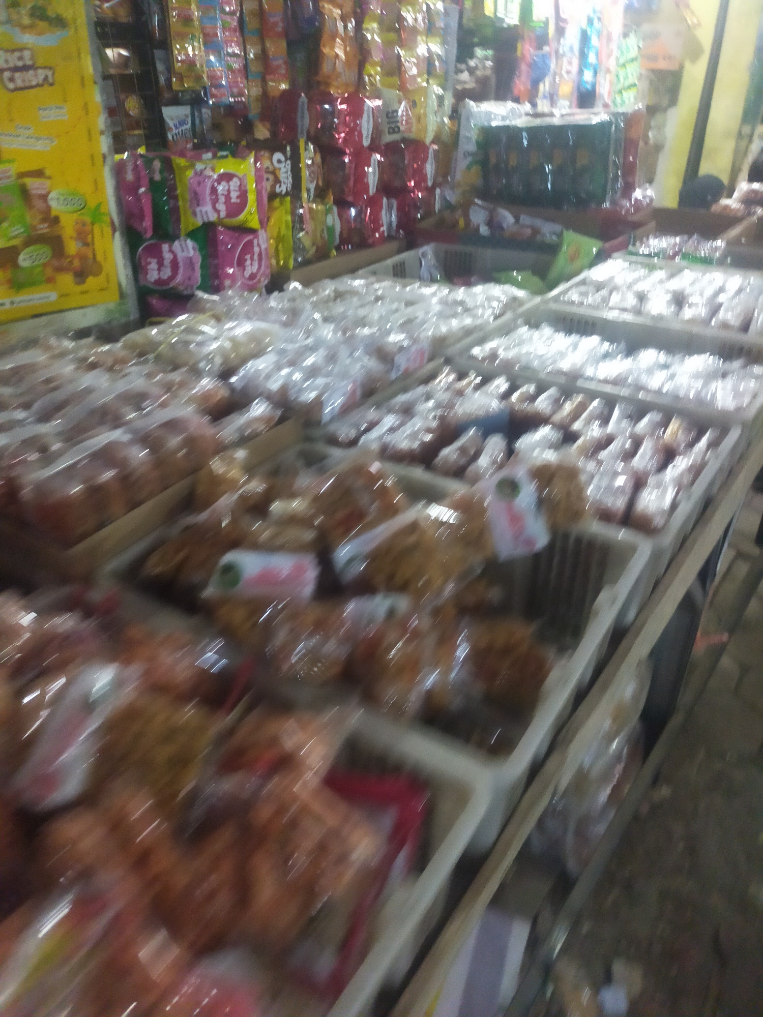 A street-level view shows a vendor stall in Trayeman, Indonesia, with numerous pre-packaged food items displayed in white plastic crates. The items appear to be snacks or sweets, with some packaging featuring pink and yellow designs. Behind the display, shelves are stocked with more packaged goods, including brightly colored bags and some larger items in dark green packaging. The overall scene is blurred and the lighting suggests it is daytime, possibly outdoors due to the rough texture of the ground visible in the lower right portion of the image.