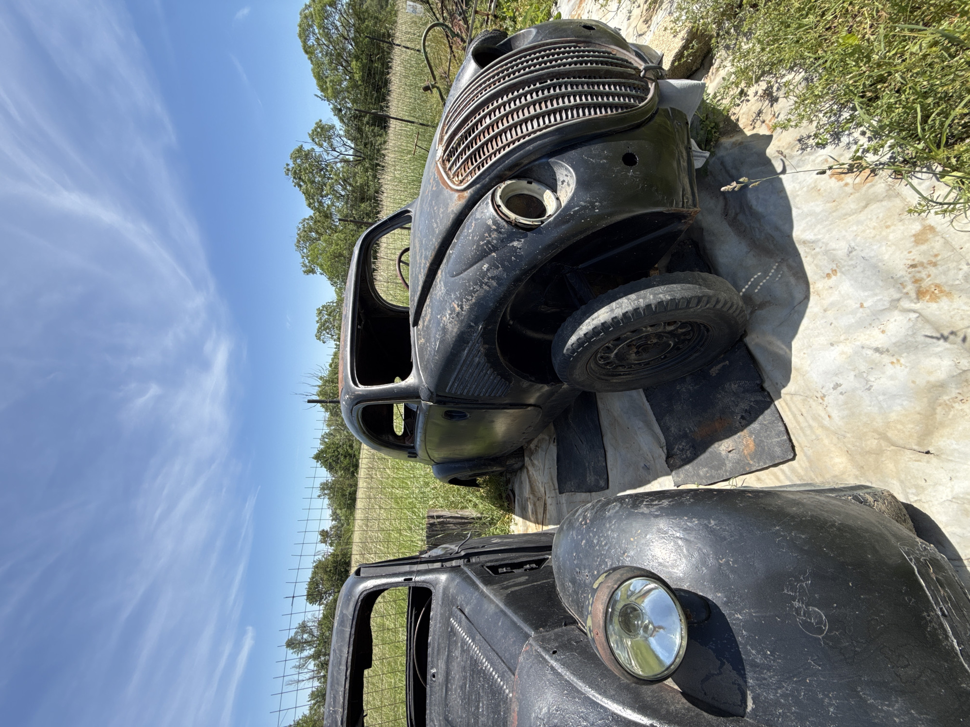 The image depicts two black, rusted car bodies in an overgrown outdoor setting under a bright blue sky with scattered clouds. The upper car body is more complete, featuring a distinctive radiator grill, a single tire, and remnants of the frame. The lower car body consists of a fender with a single headlight. The surroundings are unkempt, with overgrown grass, scattered debris, a wire fence, and trees in the background. There's no visible sign indicating the location is in the city of Omurtag, Bulgaria. No people are visible in the frame.