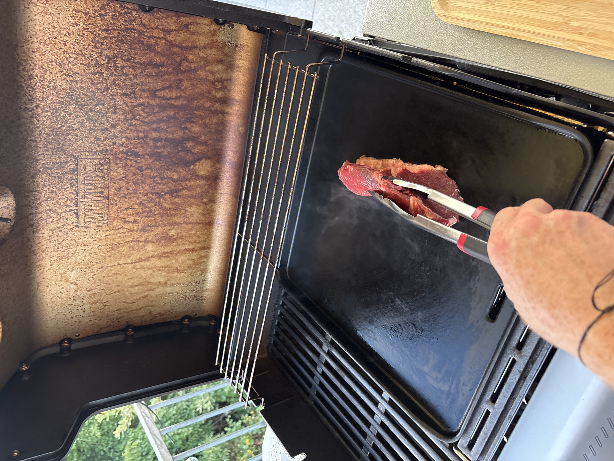 The image depicts a close-up, eye-level view of a Weber grill with its lid open. The grill has a griddle insert on the right side and a standard grate on the left. A piece of raw red meat, held by silver tongs with red grips, is being placed onto the hot griddle. Wisps of steam rise from the griddle around the meat. The interior of the grill lid is stained with grease and has a raised "Weber" logo. The scene appears to be set on a balcony or patio, with a glimpse of green foliage and a railing visible in the background. A small portion of a wooden cutting board is visible on the far right edge of the image. This scene is located in Viernheim, Germany, as indicated by the user's prompt.