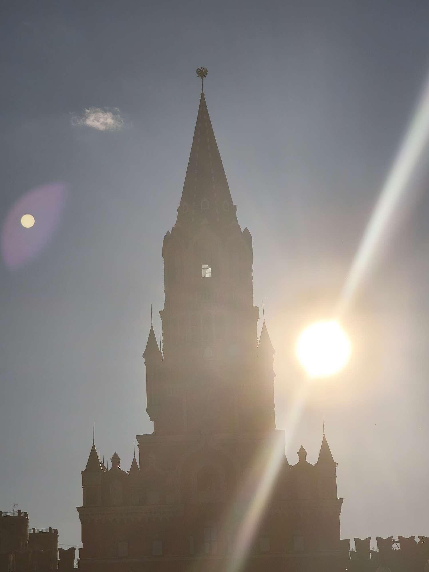This is a vertical, medium shot photograph of the Tsarevokokshayskaya Tower in Yoshkar-Ola, Russia, captured during daylight. The tower, constructed of red brick, dominates the frame with its tall, pointed spire topped by a double-headed eagle emblem. The sun is prominently positioned to the right of the tower, creating a lens flare and a bright beam of light that cuts diagonally across the sky. Another lens flare, appearing as a pale yellow circle with a purple halo, is visible to the left of the tower. A small, wispy cloud is present in the upper left quadrant of the sky. The sky is a hazy blue, with the intense sunlight causing overexposure in the central and right portions of the image. The lower part of the image shows the crenellated battlements and turrets of the tower and adjacent structures. No people are visible in the frame.