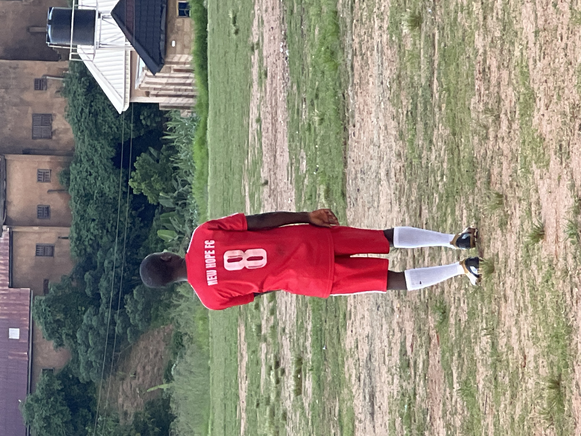 A male soccer player wearing a red jersey with "NEW HOPE FC" and the number "8" on the back stands on a dirt and grass field. He is also wearing red shorts and white socks. In the background, there are buildings, lush green vegetation, and a water tank. The location is a soccer field in Jos, Nigeria.