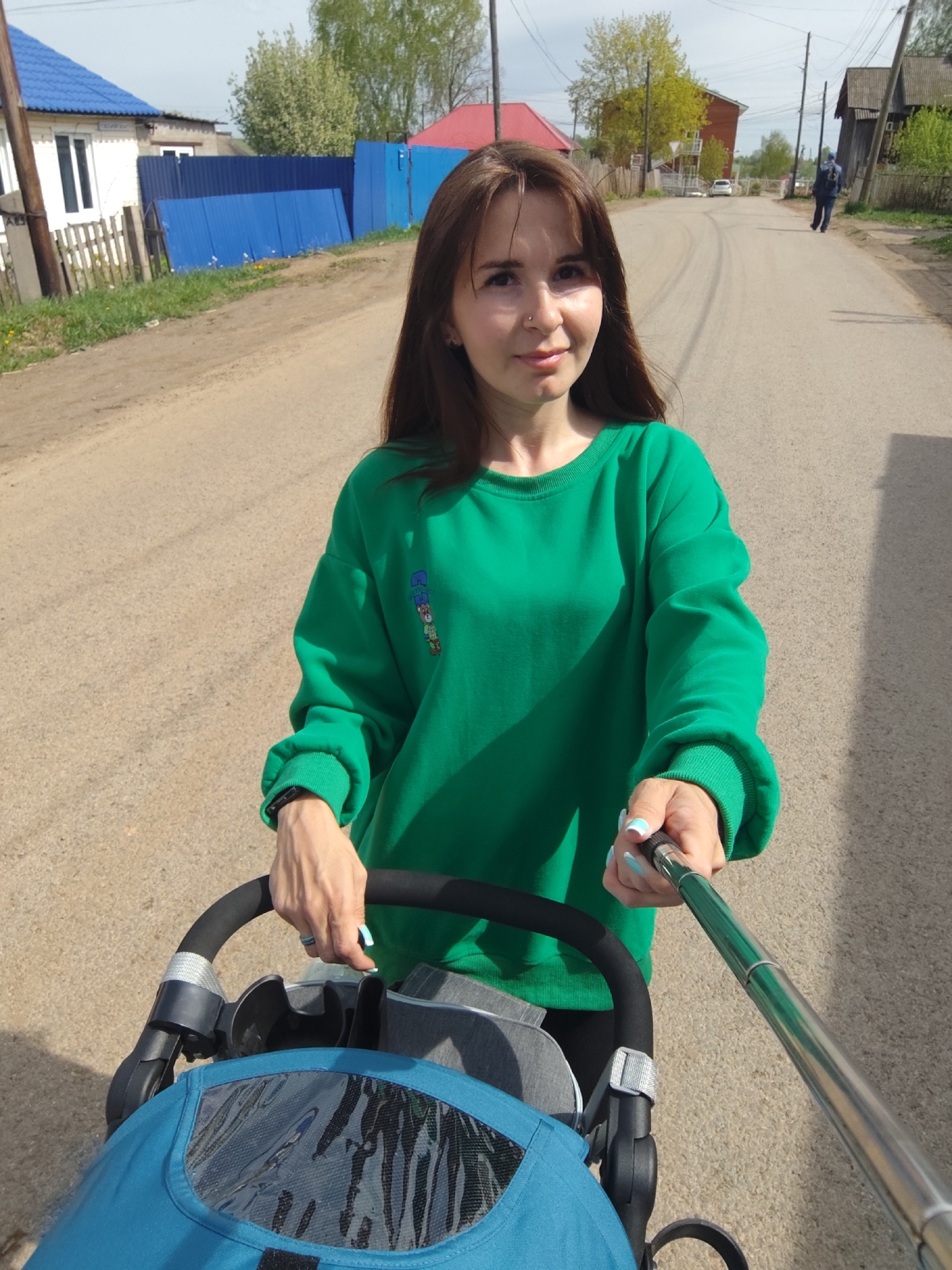 A woman in a green sweatshirt stands on a dirt road in Фоки, Russia, holding a selfie stick and pushing a blue stroller. The road stretches into the distance, where a person walks in the same direction. Buildings and fences flank the road, including a white house with a blue roof and a blue fence.