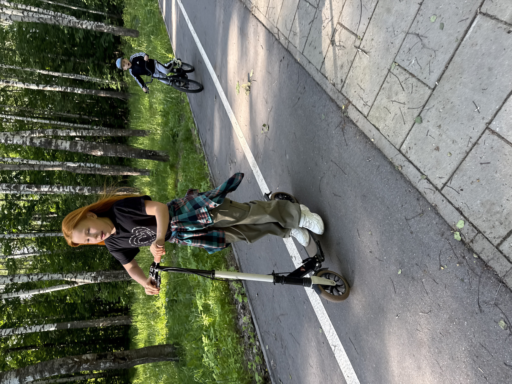 On a paved path in what appears to be a park setting in Veliky Novgorod, Russia, a young person with long red hair is riding a white and black scooter. They are wearing a black t-shirt, green pants, white sneakers, and a green and blue plaid shirt tied around their waist. The path is marked with a white line. In the background, another young person is riding a bicycle. The environment includes trees and grass. The day appears sunny.