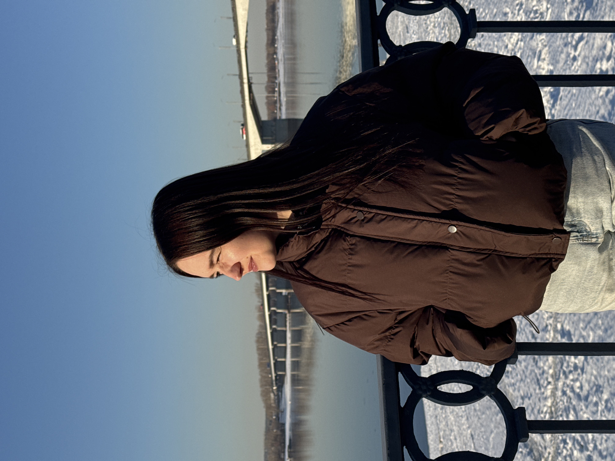 A young adult female is depicted from the waist up, positioned slightly to the right of the frame, looking towards the right. She has long, dark brown hair and fair skin, with her left cheek illuminated by direct sunlight, revealing a subtle smile. She wears a brown, puffy winter jacket with visible buttons and light blue denim jeans. The scene is located on the bank of the Kama River in Perm, Russia, identifiable by the Kommunalny Bridge (Perm Bridge) which spans the wide river in the mid-ground. The river's surface reflects the clear blue sky and shows areas of snow along its edges. Multiple vehicles are visible on the multi-span concrete bridge. In the foreground, a dark metal railing with decorative circular elements runs horizontally, separating the subject from a snow-covered embankment that descends to the river. The sky is clear and bright blue, indicating a sunny winter day.