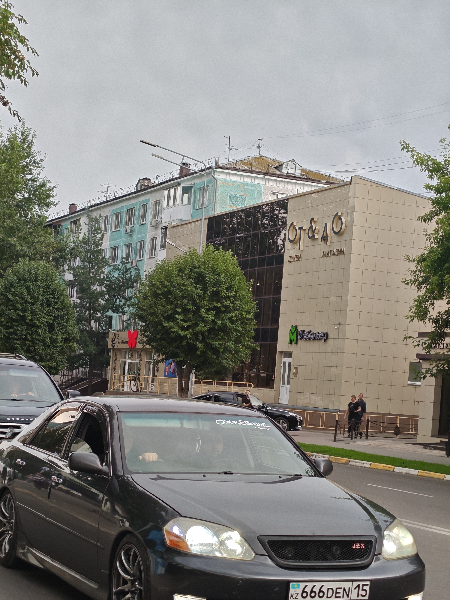 The image captures a street scene in Petropavl, Kazakhstan, featuring a dark-colored sedan in the foreground. The car is on a paved road with a yellow and white striped curb. Behind the sedan, a light-colored building with large glass windows is visible, displaying signage that reads "ОТ & ДО" and "ДУКЕН МАГАЗИН". Below this, a green logo with the text "Мебельвер" is also present. To the left of this building, a multi-story residential building with a turquoise and white facade stands. Trees line the street, with some partially obscuring the buildings. Two individuals, a man and a woman, are walking on the sidewalk in front of the light-colored building. In the background, more trees and utility poles are visible against a cloudy sky. The license plate on the foreground car is "666DEN 15" with "KZ" indicating Kazakhstan.