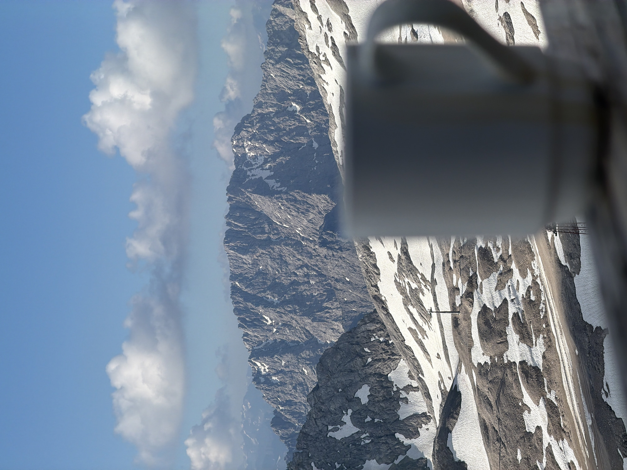 The image depicts a mountain landscape, likely in the vicinity of Garmisch-Partenkirchen, Germany, given the context of the prompt. In the foreground, a white ceramic mug is blurred, suggesting it is close to the camera. Behind the mug, the landscape unfolds with rugged, snow-dusted mountains under a bright blue sky dotted with white clouds. The terrain is characterized by rocky outcrops and patches of snow, indicating a high-altitude environment. The distant mountains are sharply defined, showcasing their craggy textures. A lone ski lift pole is visible on a slope in the mid-ground, and the presence of what appears to be a path or road suggests it may be a popular tourist or recreational area. The overall scene evokes a sense of serene, crisp mountain air and a peaceful morning.