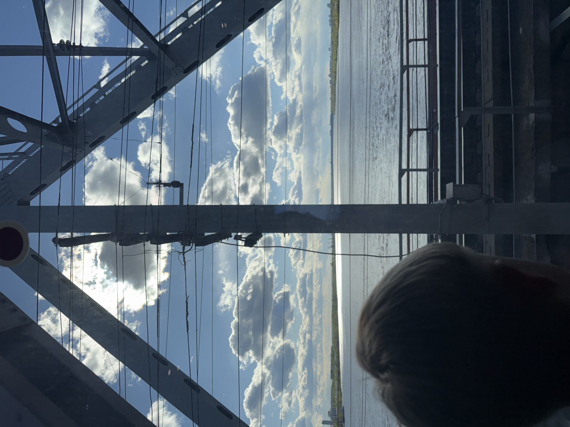 The image shows a view from the Kama Bridge in Perm, Russia. The steel framework of the bridge is visible on the left side of the frame. A body of water with a distant shoreline occupies the center of the image. The sky is blue with fluffy white clouds. A person's head is visible in the lower right corner of the frame. Electrical wires run along the left side of the image.
