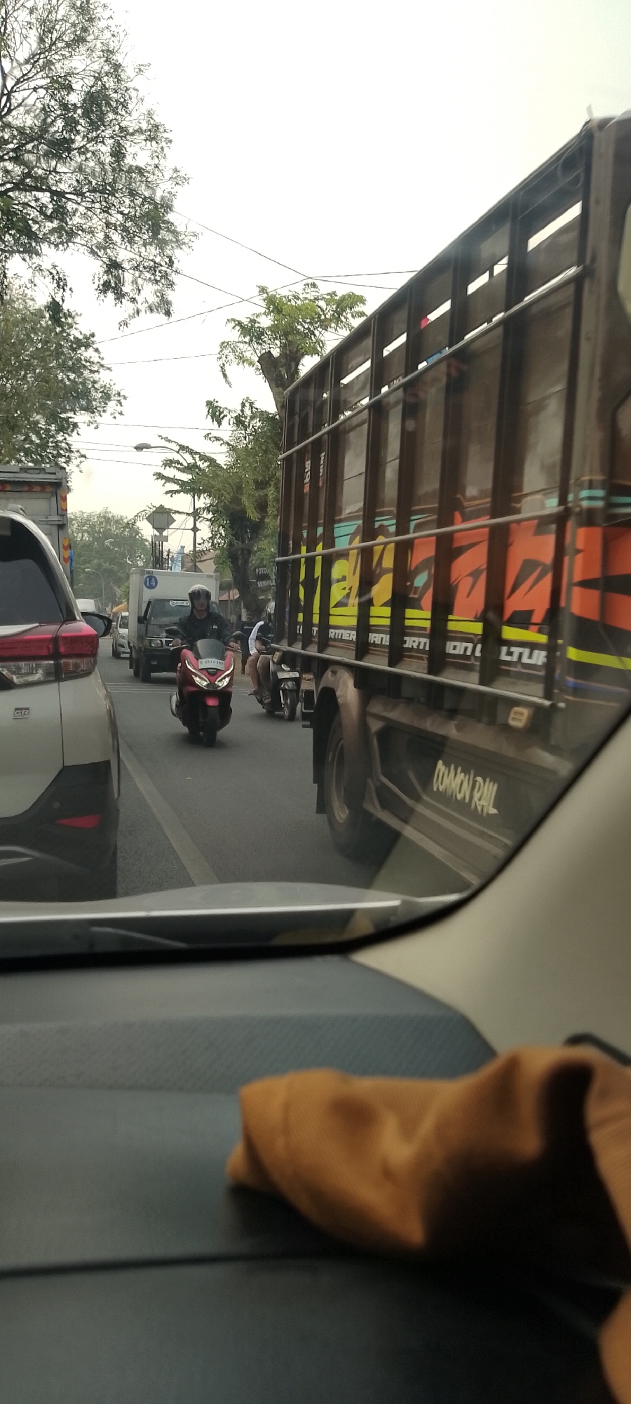 A first-person perspective from inside a vehicle shows a traffic jam on a city street. The sky is overcast. In the foreground, the dashboard and windshield of the car are visible, with a brown cloth placed on the dashboard. In the middle ground, a line of vehicles is stopped on a paved road. A white SUV is on the left, followed by a red scooter with a rider wearing a helmet, then a white truck, and a black truck with a large wooden cargo bed. The black truck has colorful graphics on its side and the words "COMMON RAIL" are visible. Several other motorcycles are also in the traffic. Trees and utility poles line the sides of the road. The scene appears to be in Panjang, Indonesia, a port city known for its industrial and commercial activities.
