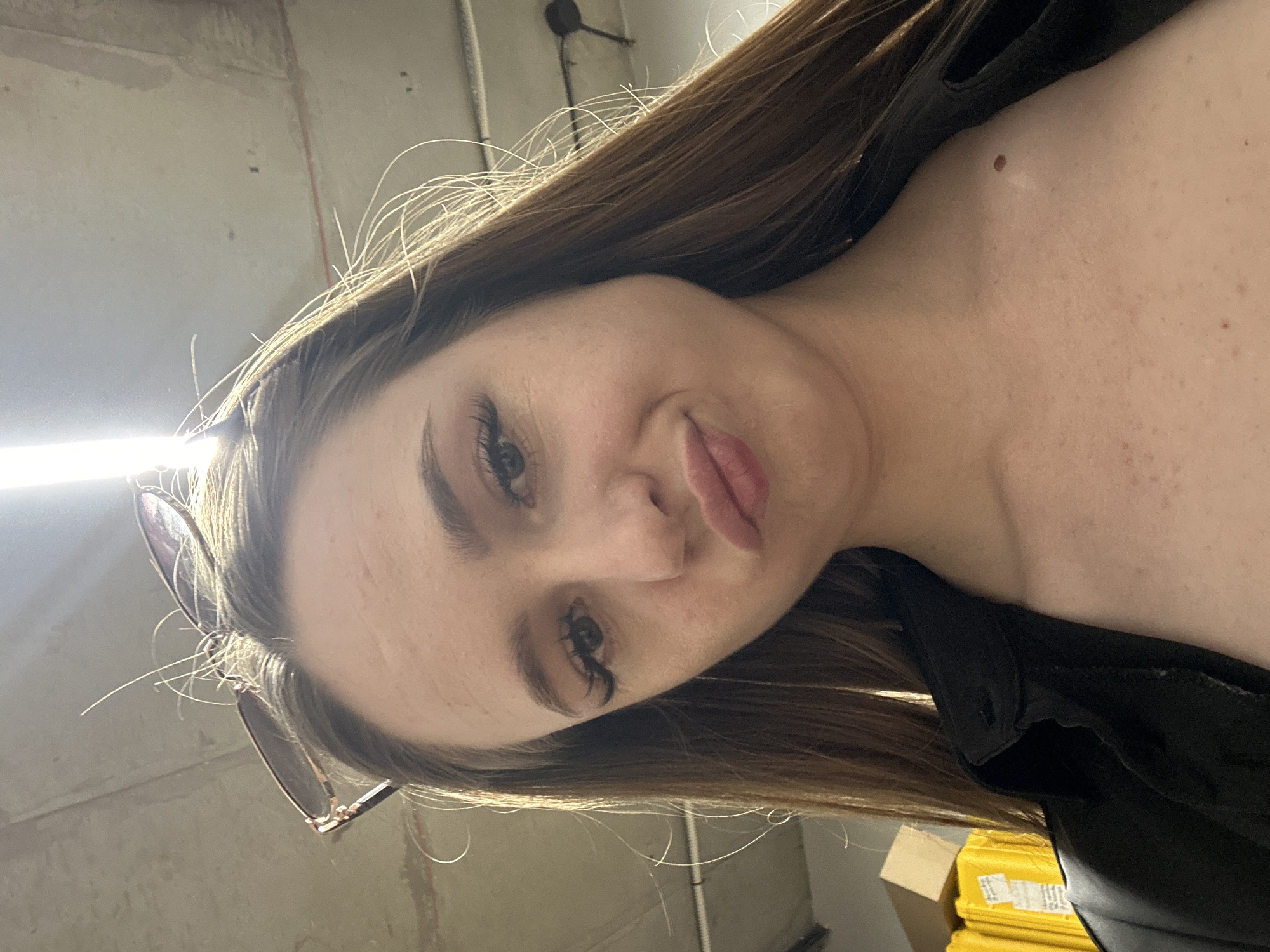 A young adult female is captured in a low-angle, close-up selfie, her face framed by long, brown hair that is partially obscured by sunglasses perched on her head. She has light skin, dark eyebrows, and long eyelashes. Her lips are pursed in a pout, and she appears to be looking directly at the camera. She is wearing a black collared shirt. The background is indistinct, showing a concrete ceiling with lighting fixtures and some yellow and brown boxes. The overall setting appears to be indoors, possibly a warehouse or industrial space.