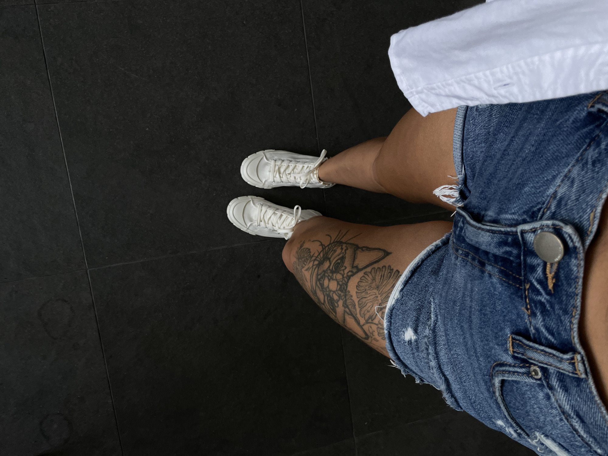 In Kediri, Indonesia, a person stands on dark tiled flooring. They are wearing ripped denim shorts and white sneakers. A tattoo is visible on their left thigh. A white garment, possibly a shirt or blouse, is draped on their right leg. The scene is viewed from a high angle, looking down.