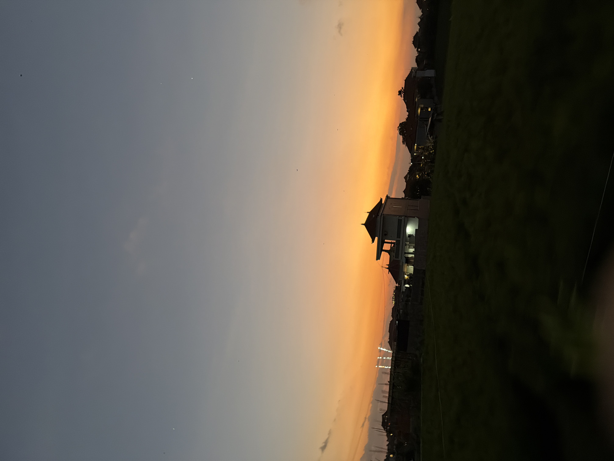 The image depicts an outdoor scene at dusk or dawn in North Kuta, Indonesia, characterized by a vibrant gradient sky, architectural structures, and undeveloped land.
The sky occupies approximately the upper two-thirds of the frame, transitioning horizontally and vertically. The lower sky exhibits a bright orange-yellow hue along the horizon, fading upwards into a soft peach, then a pale blue-grey, and finally a darker blue-grey at the top. A distinct, thin crescent moon is visible in the upper-left quadrant of the sky. Several faint, small, white pinpricks, consistent with stars or distant celestial bodies, are scattered across the darker upper sky. A few wispy, elongated cloud formations are present near the horizon on the left side, partially obscuring the bright orange light. Scattered throughout the sky are multiple tiny, dark specks, likely small birds in flight.
The midground features a prominent multi-story building, approximately centered horizontally, with a distinctive dark-tiled, multi-tiered roof, indicative of traditional Balinese architecture. The building's facade is illuminated by internal or external lights, casting a warm glow and revealing a light-colored exterior. To the left of this central building, a cluster of vertical white light structures, resembling stadium or construction lights, projects upwards from beyond a dark, indistinct area. Along the entire horizon line, numerous other residential structures with traditional roofs are visible, largely silhouetted against the brighter sky. These buildings suggest a developing residential area.
The foreground, occupying the lower third of the image, consists of a dark, undifferentiated expanse of land, consistent with a field or grassy area. The color is a deep olive-green to near-black, suggesting low light conditions or dense vegetation. No distinct features or objects are discernible within this foreground. There are no identifiable people or animals visible anywhere in the image.