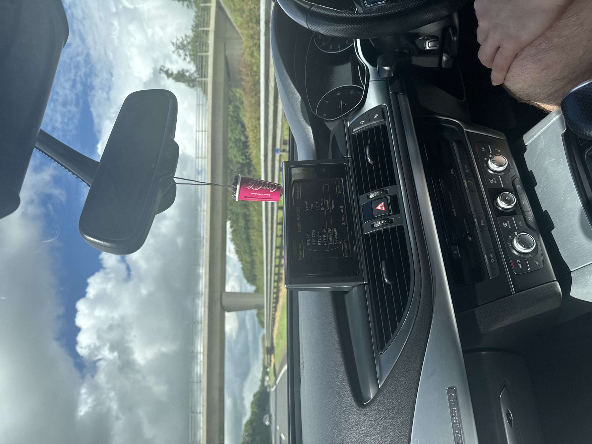 This media file captures the interior of a car driving on a highway. The perspective is from the driver's seat, looking forward. Visible subjects include the dashboard with its infotainment system displaying radio stations and settings, a rearview mirror, and the steering wheel. A person's arm and hand are visible, suggesting the driver.
The scene is set on a highway, indicated by the road, guardrails, and overpasses visible through the windshield. The weather appears to be partly cloudy, with a mix of blue sky and white clouds. The lighting suggests it is daytime, likely late morning or early afternoon.
A notable detail is a Coca-Cola branded air freshener hanging from the rearview mirror. The infotainment screen displays several radio station presets, including RTE R1, RTE 2FM, RTElyric, RTE RnaG, Today FM, and Newstalk, all common in Ireland. The time displayed on the screen is 3:18. The dashboard also shows climate control settings and other vehicle functions. The overall impression is a mundane journey on a highway, with the driver presumably engaged in listening to the radio.