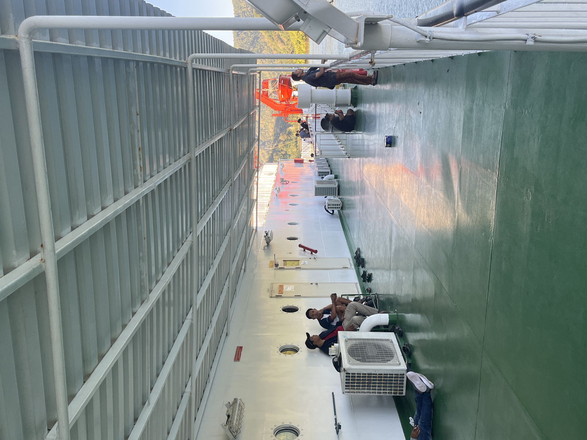 The image depicts the deck of a ferryboat, likely in the vicinity of Batas Desa Skotong Timur, Indonesia, given the lush green hills visible in the background. The deck is a light green color and is partially covered by a white metal canopy. Several people are present on the deck, some sitting and conversing, others standing and looking out at the water.
In the foreground, a person lies on their side, resting. To the left, near a white air conditioning unit, two men are seated, one wearing a black baseball cap and a red jacket, the other in a dark t-shirt and shorts. Further down the deck, several pairs of sandals are neatly placed on the floor.
Mid-deck, a red lifeboat davit system is visible. A person in a dark shirt and patterned pants stands near the railing, looking out towards the water and the distant hills. Other individuals are seen scattered on the deck, some appearing to be waiting or relaxing. The water is calm and reflects the light from the sky. The overall scene suggests a relaxed atmosphere, typical of ferry travel.