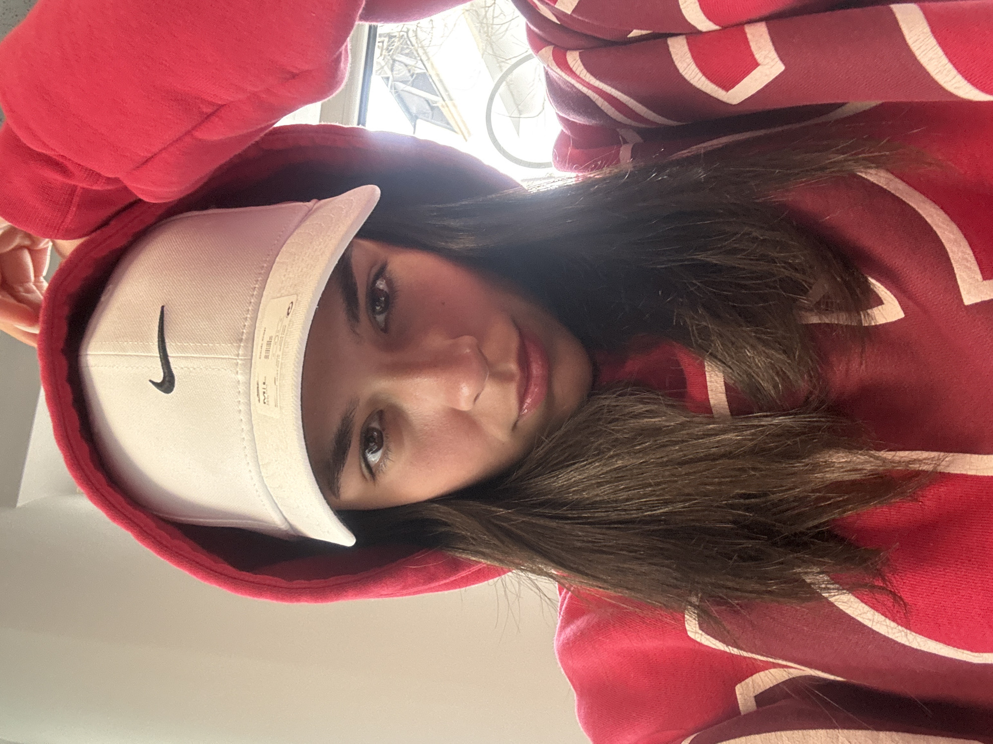 A young woman is posing for a selfie. She is wearing a red hooded sweatshirt and a white baseball cap with a black Nike logo on the side. Her long dark hair is visible under the cap. The background appears to be indoors, with a window visible in the upper left corner. The image is slightly rotated. The location is not Gabrovo, Bulgaria.