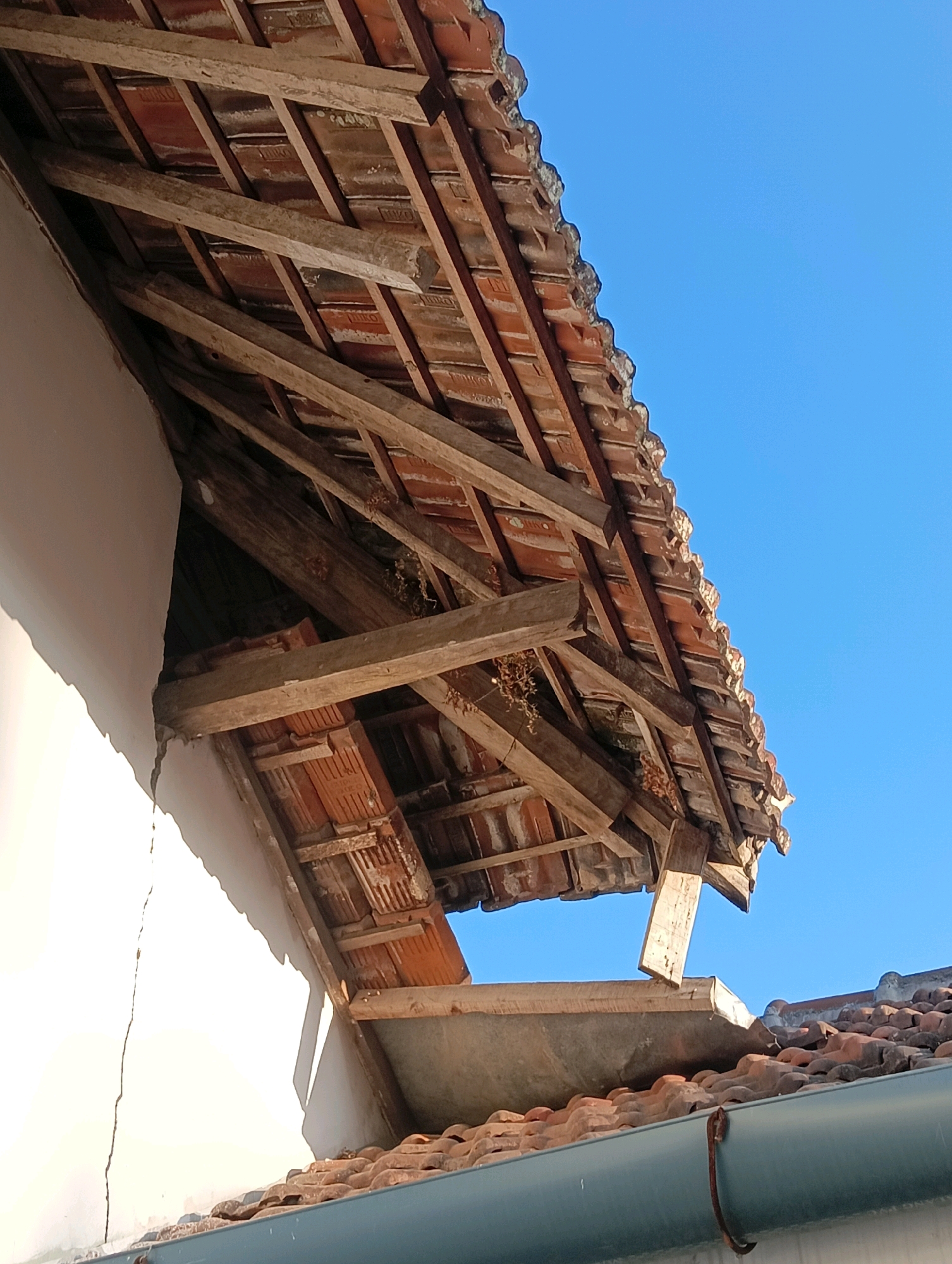 The image shows a damaged roof made of terracotta tiles and wooden beams. The roof appears to be part of a building under repair or in a state of disrepair. The wooden structure supporting the tiles is visible, with some beams appearing broken or displaced. A white wall of an adjacent structure is also visible, showing a crack running vertically. The sky is clear and blue. The scene is likely in Sukosari, Indonesia, given the context provided. No people are visible in the image.