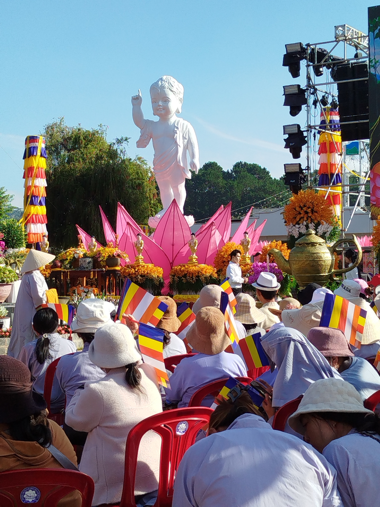 The image depicts a Buddhist celebration in Da Lat, Vietnam. A large white statue of a child Buddha, with its right arm raised, stands atop a display of pink lotus flowers. In front of the statue, rows of people sit in red plastic chairs, many holding Buddhist flags. They wear various hats and light-colored clothing. To the left of the statue is a tall, colorful pillar. To the right, stage equipment and decorations are visible. The sky is clear and blue.