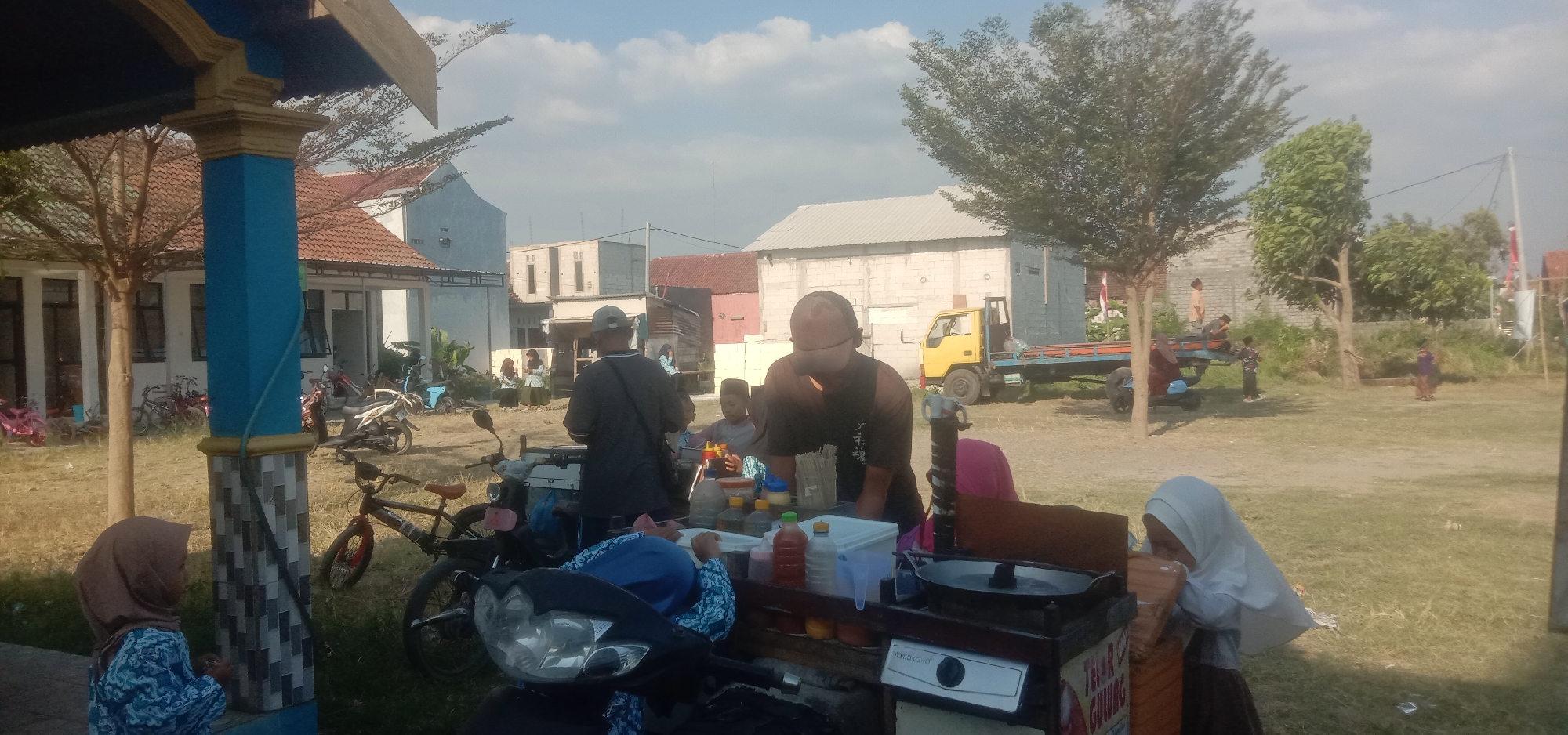 The image depicts an outdoor scene in Cangkringrandu, Indonesia, likely a street or market area. In the foreground, a young girl wearing a light blue and white patterned shirt and a brown hijab is standing next to a blue and gold decorated pillar, looking away from the camera. To her right, a food cart is set up on a motorcycle. On the cart, there's a gas stove with a frying pan, various bottles of condiments, and other food preparation items. A person wearing a white hijab is partially visible on the right side of the cart, appearing to be interacting with the cart.
Further back, several people are gathered around another food stall. A man in a dark t-shirt and a cap is standing, facing away from the camera, seemingly serving food. Another person, a child, is seated at the stall. Behind them, a yellow truck with a flatbed trailer is parked. In the background, various buildings with tiled roofs and some structures with corrugated metal roofs are visible, suggesting a semi-urban or rural environment. Several motorcycles and bicycles are parked in the area, indicating a common mode of transportation. The ground is covered in dry grass and dirt, and trees are scattered throughout the scene. The sky is partly cloudy.