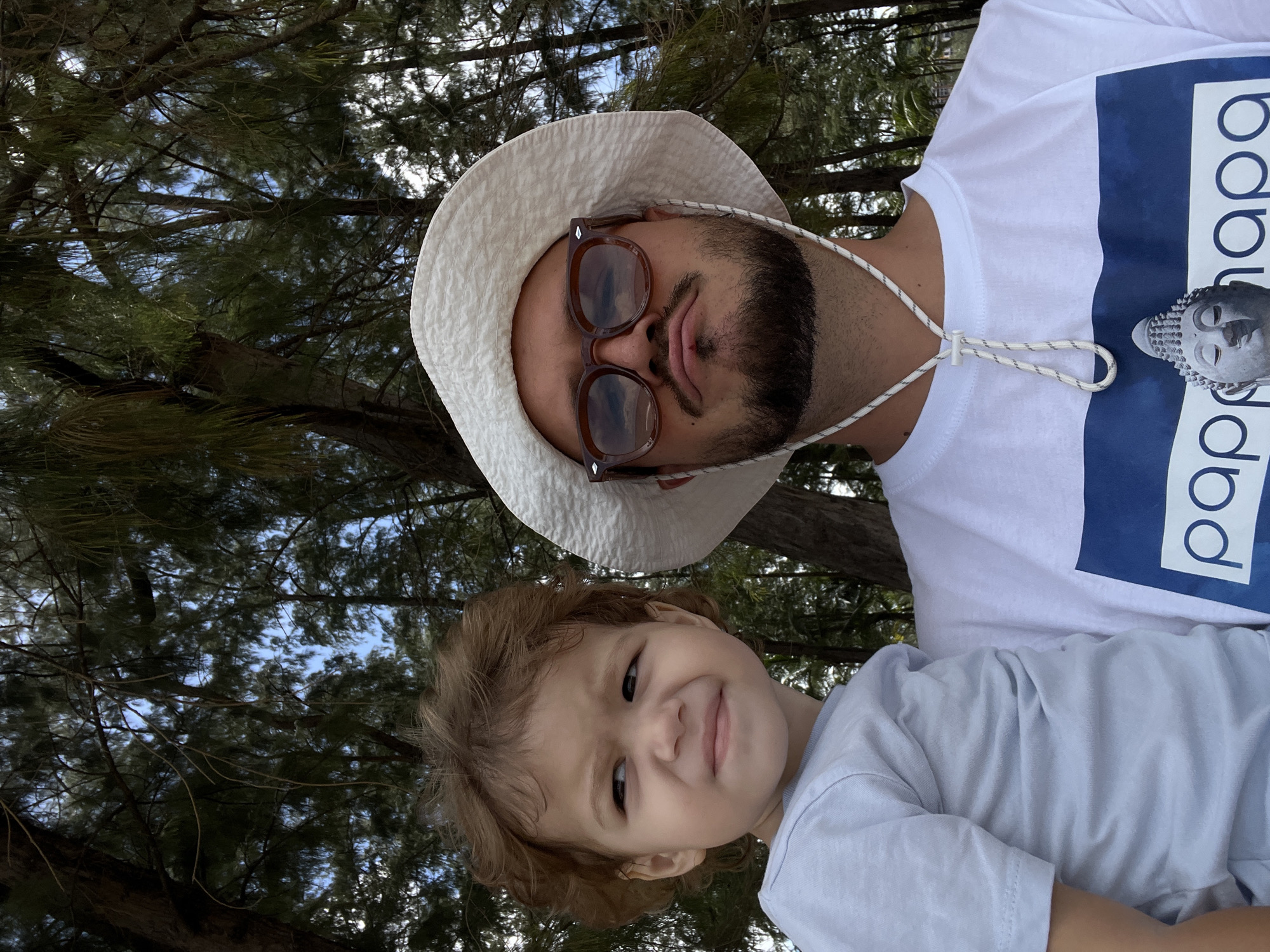The media file is a selfie taken from a low angle, featuring a man and a young child in an outdoor setting with trees and blue sky visible in the background. The man, wearing a white bucket hat and sunglasses, is positioned in the upper portion of the frame. He has a beard and is looking downwards with his lips slightly pursed. He is wearing a white t-shirt with a large blue graphic on it, which appears to have text. A white cord is attached to his hat and goes down the front of his shirt. The child, positioned in the lower portion of the frame, is looking up at the camera with a slight smile. The child has light brown curly hair and is wearing a light blue t-shirt. The overall impression is one of a casual, relaxed moment, possibly during daylight hours given the bright sky. The presence of trees suggests a natural or park-like environment. The text on the man's shirt is partially visible and appears to be in a sans-serif font.