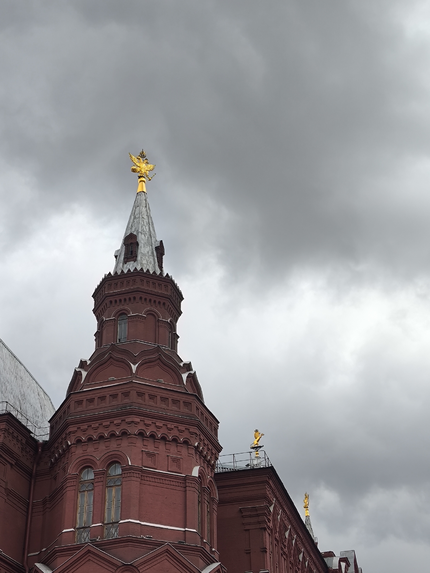 The image features a close-up view of a majestic, deep red brick building, characterized by its intricate historical architecture. A prominent octagonal tower rises in the foreground, topped with a tall, pointed spire covered in grey, scale-like material. Crowning the very peak of the spire is a magnificent golden sculpture of a double-headed eagle, a recognizable national emblem of Russia.
Further along the building's roofline to the right, additional smaller golden decorative elements, also appearing to be stylized eagles or similar heraldic figures, are visible. The building's facade is adorned with decorative cornices, stepped details, and arched windows, typical of late 19th-century Russian revivalist style.
The sky is heavily overcast with thick, dark grey clouds, indicating a gloomy day. The dramatic cloud cover fills most of the background, creating a stark contrast with the warm red of the building and the gleaming golden accents. There are no people or specific activities depicted. The scene suggests a daytime setting, albeit under very muted natural light. Given the golden double-headed eagle and the architectural style, the building is likely a significant historical landmark in Moscow, Russia, such as the State Historical Museum. No text is visible in the image.