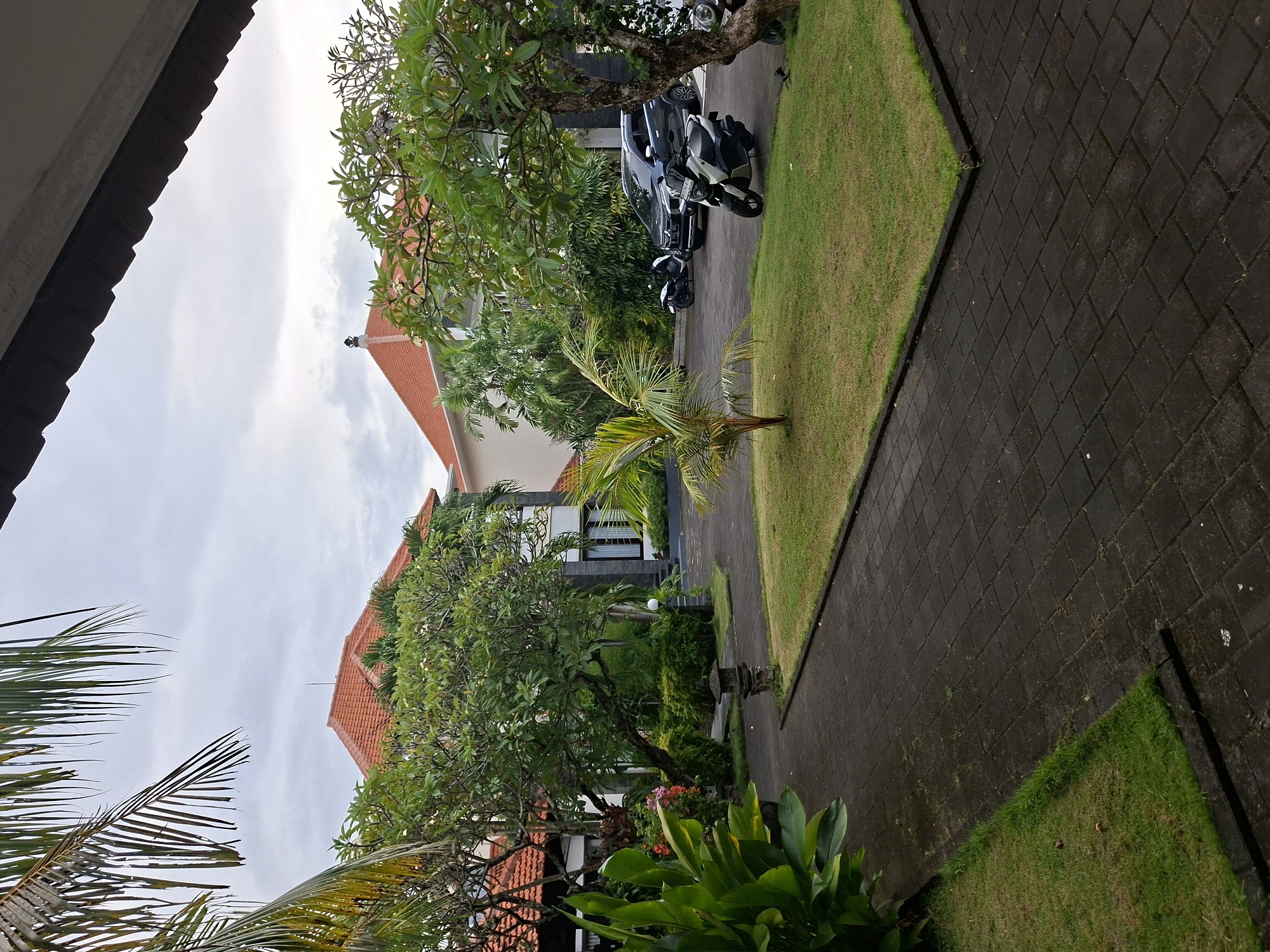 This image depicts a tranquil outdoor scene, likely within a residential area or resort in Kuta Selatan, Indonesia. The overall setting is characterized by lush tropical vegetation, traditional architecture, and well-maintained grounds under an overcast sky.
In the foreground and midground, a dark, paved pathway made of square interlocking stones winds through the scene, bordered by neatly trimmed grassy lawns. Abundant greenery dominates the view, including several palm trees, frangipani trees with sparse white blossoms, and various other broad-leafed tropical plants.
Houses with distinctive red-orange tiled roofs are visible, partially framed by the dense foliage. One house in the midground showcases modern architectural elements with large dark-framed windows set against light-colored walls. A paved driveway leads to a parking area where a dark-colored SUV or car is parked alongside two scooters or motorcycles, one dark and one lighter in color. A traditional Balinese-style stone offering stand is subtly placed among the plants, serving as a distinct cultural indicator.
The sky is uniformly overcast and grey, suggesting a cloudy daytime, possibly late morning or afternoon, as there are no sharp shadows cast by direct sunlight. No people are visible, and no specific activity or event is taking place, presenting a static and peaceful snapshot of the environment. There is no visible text in the image.