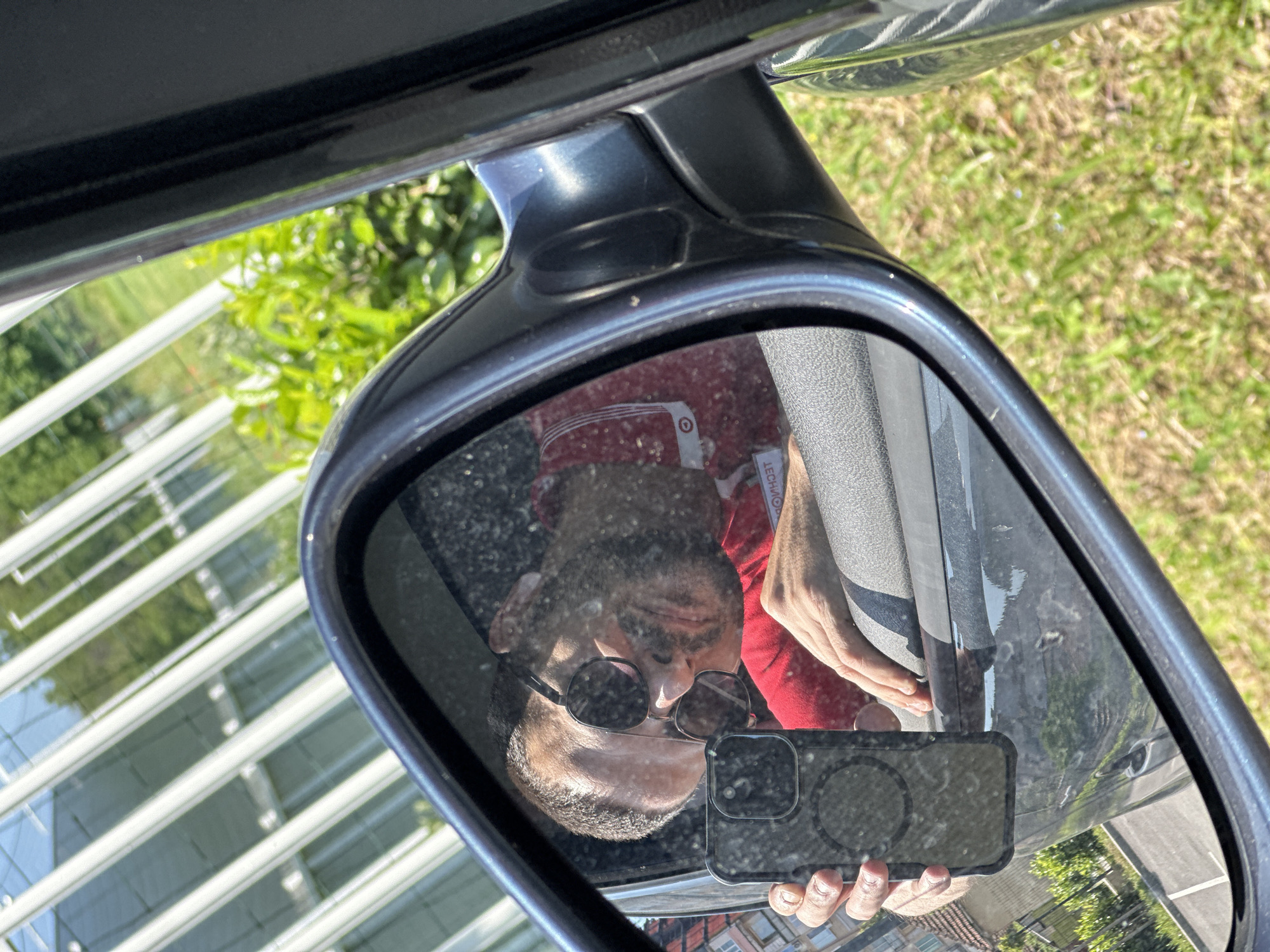 The image shows a reflection in a car's side mirror. A person, presumably taking the photo, is visible holding a smartphone and wearing sunglasses and a red and white shirt. The reflection also shows part of the car's door and potentially a sleeve. Outside the car, the background shows greenery, suggesting a park or grassy area. Based on elements visible beyond the mirror's frame, there is a modern building with glass facade structure. Without specific details to link the structures in the image to locations within Lovech, it is difficult to verify that the picture was taken in that location.