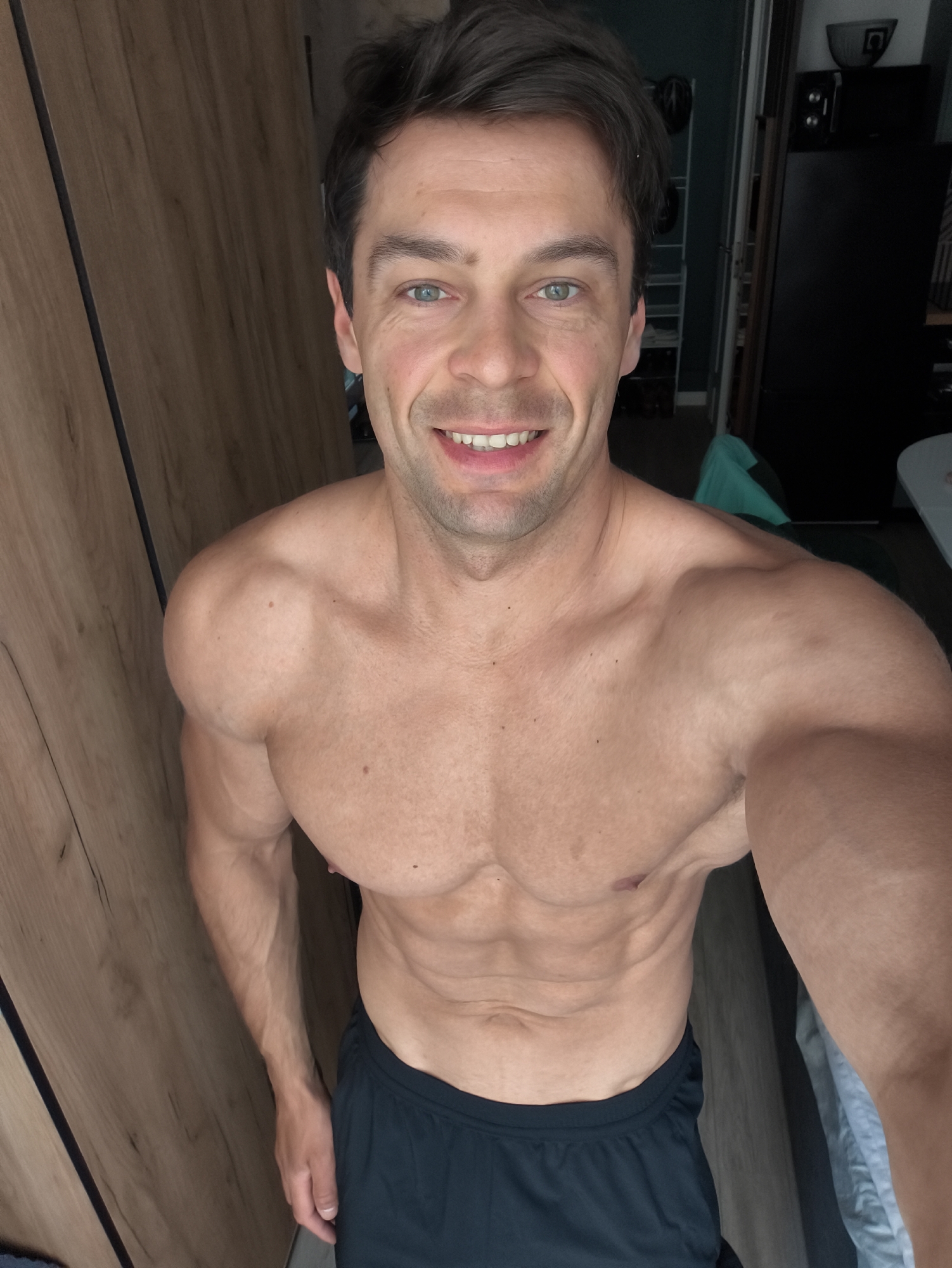 A man is taking a selfie in a room. He has fair skin, short brown hair, and blue eyes. He is shirtless, showing his toned physique, and is wearing dark shorts. He is smiling at the camera. The background shows a wooden closet on the left and a dark refrigerator and some shelves on the right. The lighting appears to be artificial, coming from the front. There is no indication that this image was taken in Moscow, Russia.
