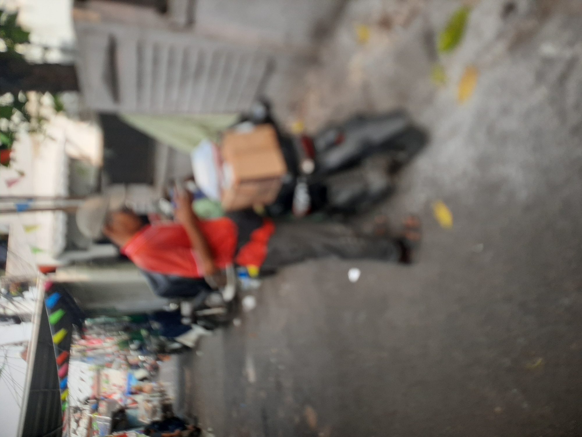 In Central Jakarta, Indonesia, an out-of-focus image shows a person wearing a red shirt and a cap standing next to a motorcycle. The motorcycle has a cardboard box strapped to the back. The person appears to be looking at their phone. In the background, there are other motorcycles parked along a street lined with buildings and colorful flags. The scene is somewhat cluttered, with what appear to be scattered leaves and debris on the ground. The overall impression is of a busy urban environment.