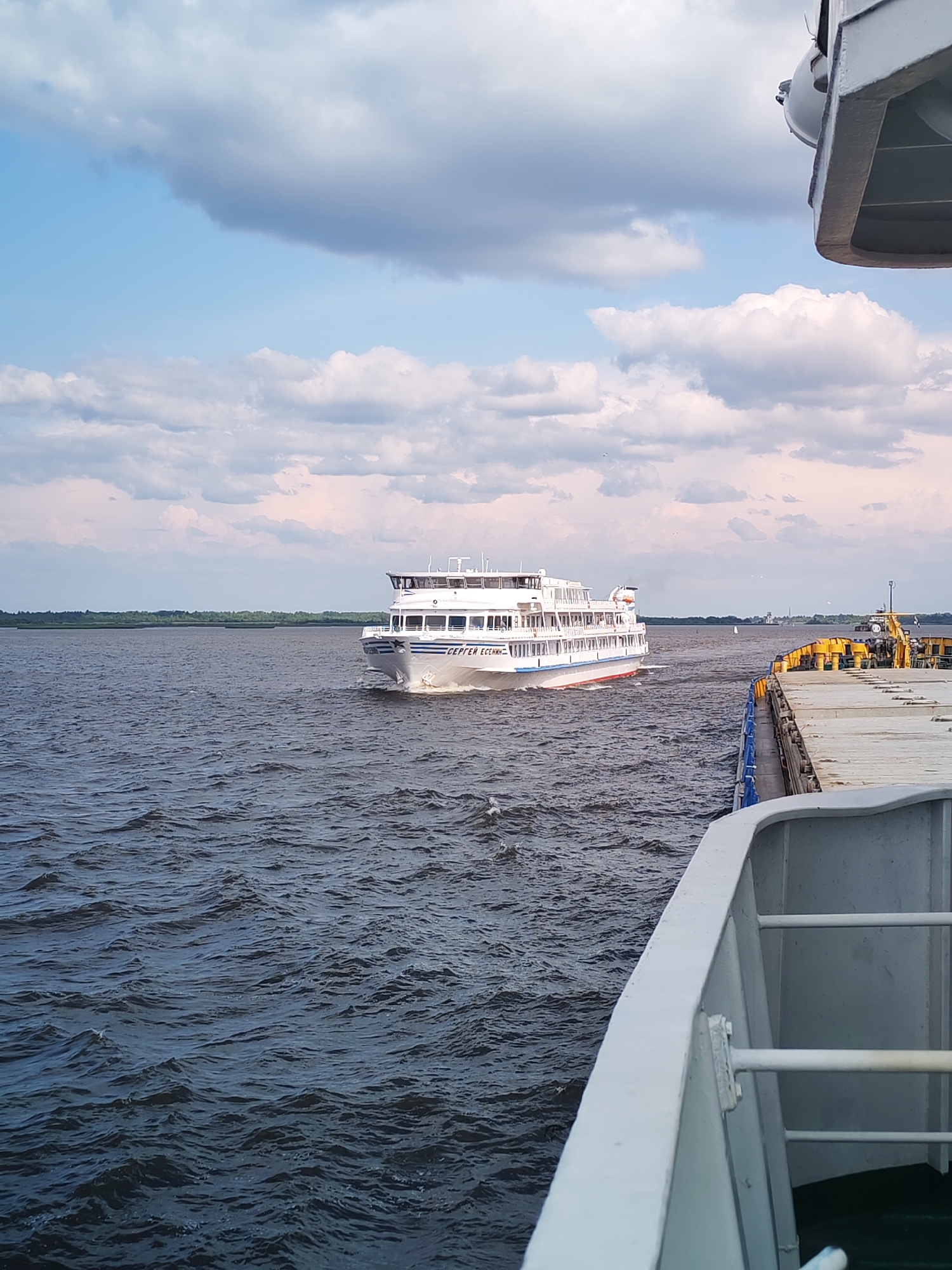 A white passenger ship, identified by the Cyrillic text "СЕРГЕЙ ЕСЕНИН" (Sergei Yesenin) on its hull, is sailing on a dark, wavy body of water. The ship has multiple decks with numerous windows. The sky above is partly cloudy with patches of blue. In the foreground, to the right, the edge of another vessel is visible, painted white with a blue railing. Further in the distance, a green, tree-lined shore is apparent. The scene is set on the Volga River near the historically significant, partially submerged village of Krokhino, Russia, known for its abandoned St. Nicholas Church, which is not visible in this frame. The overall impression is one of maritime travel on a cloudy day.