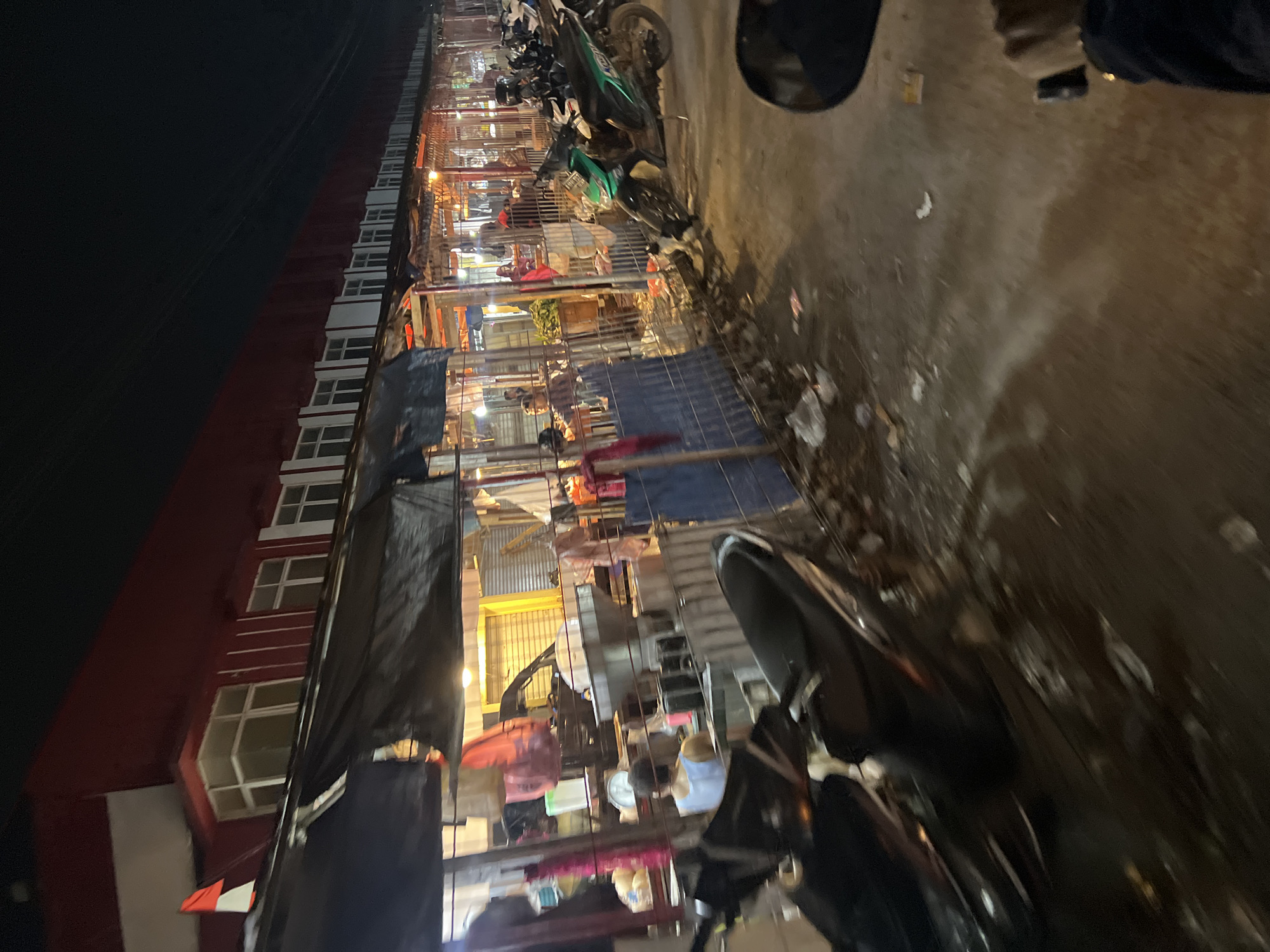 This is a night-time, low-angle, exterior shot of a market in Citeureup, Indonesia, likely from the perspective of someone on a motorcycle, indicated by the blurred handlebars and mirror in the foreground. The market appears to be a row of food stalls with awnings made of black tarpaulin. A red building with white-trimmed windows forms the backdrop. The stalls are illuminated by warm, yellow lights, casting a glow on the goods and people within. Several motorcycles are parked in front of the stalls, suggesting a busy evening. There are people in the stalls, some appearing to be vendors and others customers. The ground is paved and somewhat littered with debris. The overall impression is of a lively local market scene at night.