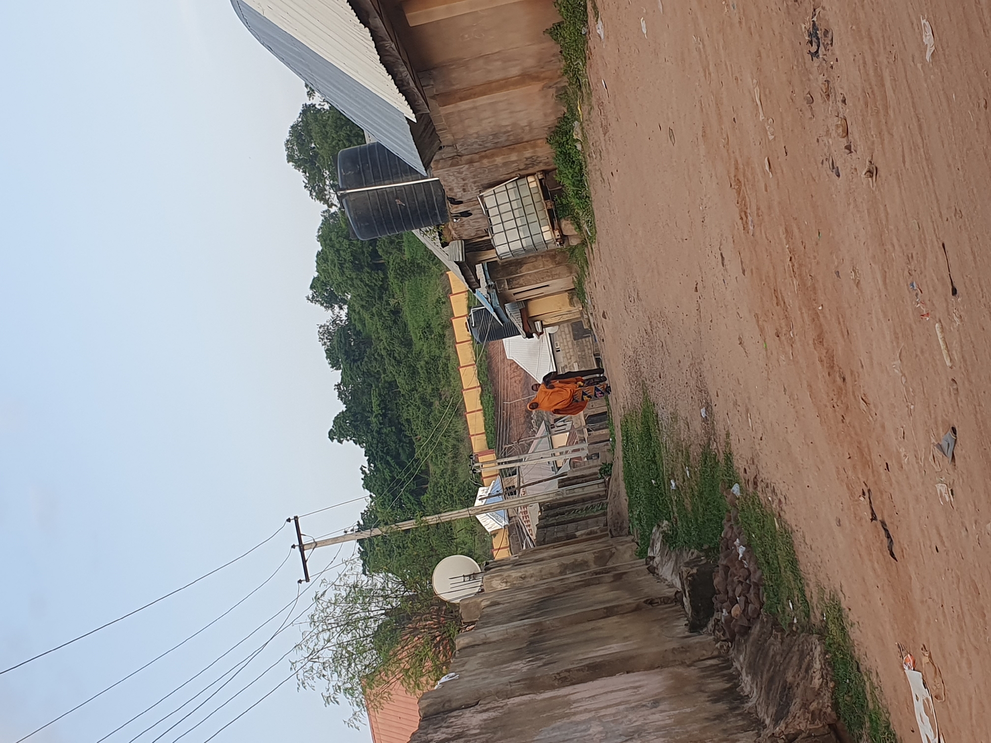 The image depicts a dirt road in a residential area of Lokoja, Nigeria. The road is flanked by concrete walls on the left and a row of buildings on the right. The buildings have corrugated metal roofs and some have large water tanks installed. In the background, a lush green hillside rises, dotted with trees and smaller structures. Overhead, power lines crisscross the sky, supported by wooden utility poles. Two individuals are walking away from the camera on the dirt road. One person is wearing an orange garment, and the other is wearing dark clothing. The environment appears to be a typical urban or peri-urban settlement, characterized by its unpaved roads, modest buildings, and the presence of essential utilities like electricity and water storage.