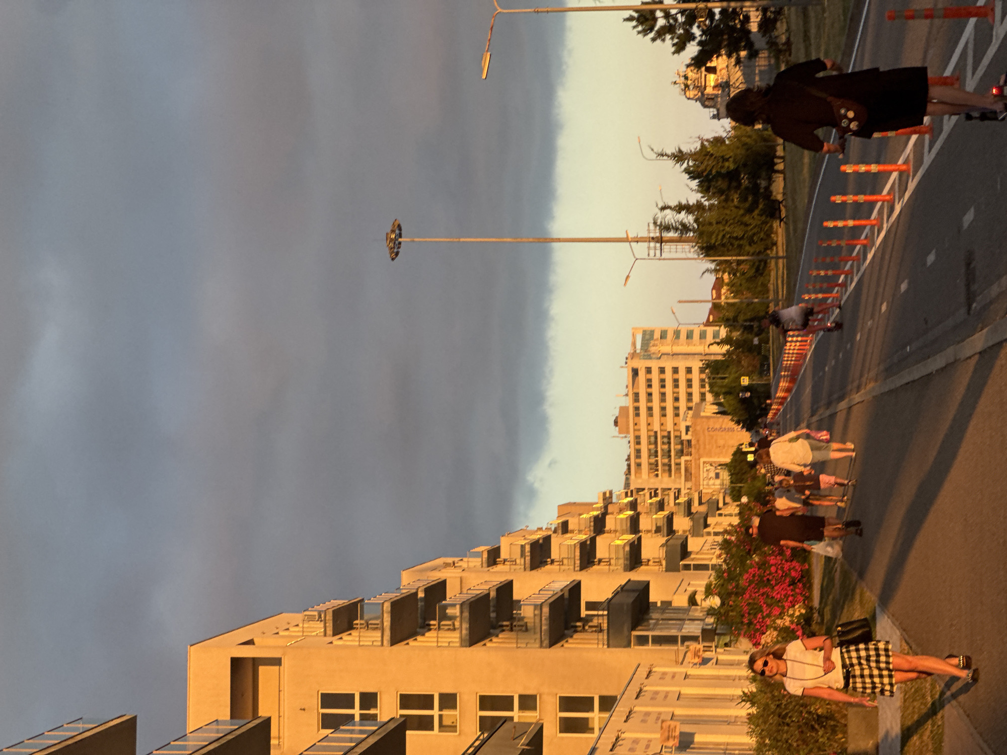 This media file depicts a scene at dusk in Sirius, Russia. The image is oriented vertically, with the sky occupying the upper portion and a street with buildings lining one side in the lower portion.
The sky is a gradient of colors, with dark, cloudy tones on the left transitioning to a lighter, warmer hue on the right, suggesting the sun is setting or has recently set. A tall, industrial-looking light pole stands prominently in the sky.
On the right side of the image, a paved road is visible, bordered by orange traffic cones and a sidewalk. Several people are walking along the sidewalk. One person in the foreground is wearing a white t-shirt and a black and white checkered skirt, holding a black purse, and appears to be looking towards the camera. Further down the sidewalk, other individuals are seen walking, some in groups.
The left side of the image features a modern architectural style with stepped buildings made of light-colored material. These buildings have many windows and outdoor units on their rooftops. Lush pink flowers are visible in a garden area between the sidewalk and the buildings.
The overall atmosphere is one of an urban evening stroll. The lighting suggests it is late afternoon or early evening, with the warm light casting long shadows. The weather appears calm. There is no visible text in the image.