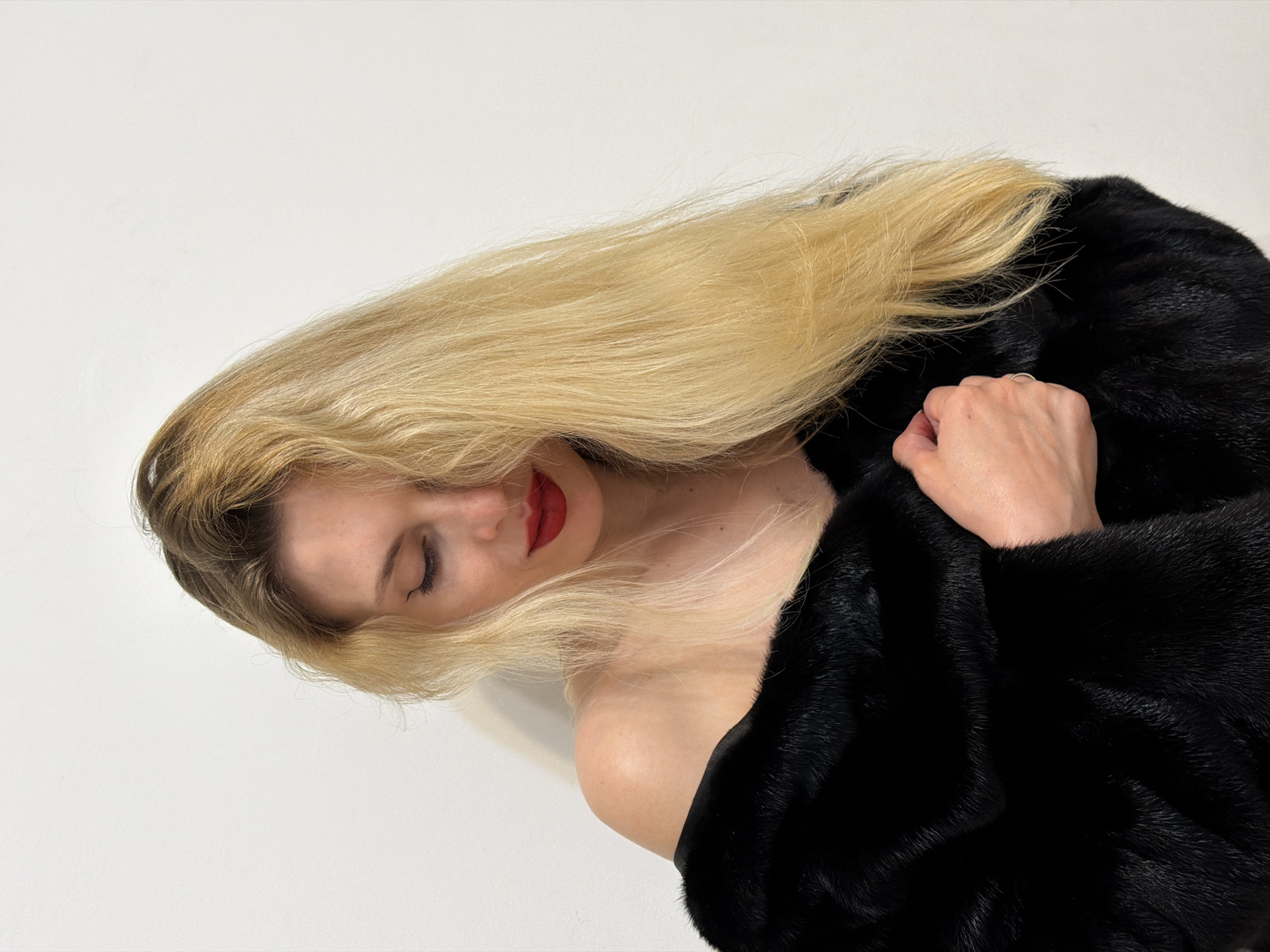 A woman with blonde hair, wearing a black fur coat, is depicted in a studio setting. Her eyes are closed and she is wearing red lipstick. Her hair is neatly styled. Her hand is visible on the coat. The background is a plain white wall. The location (Budva, Montenegro) is not evident from the image.